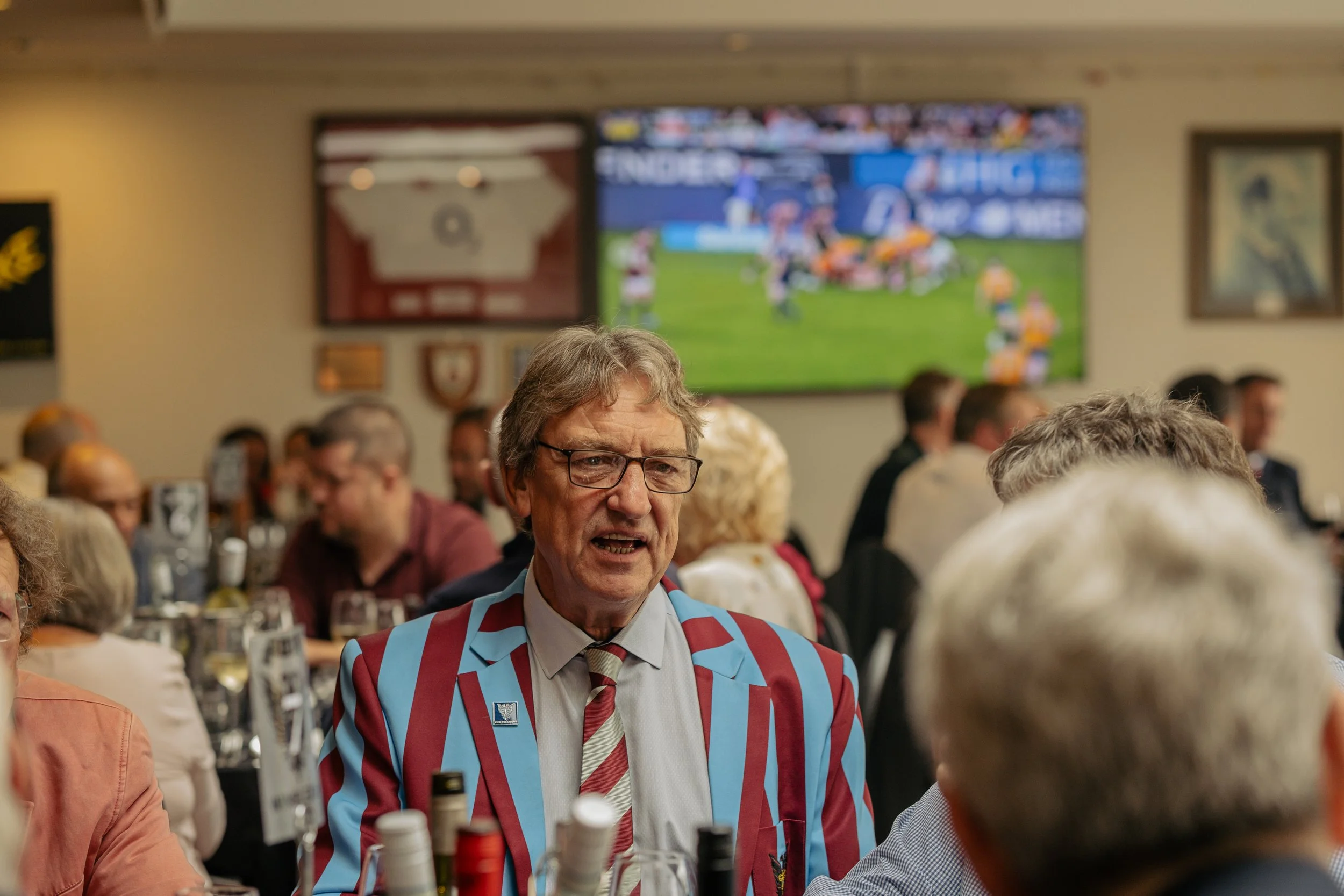 A man in a striped blazer and glasses speaking at a social gathering or event with other people, with a television and sports game in the background.