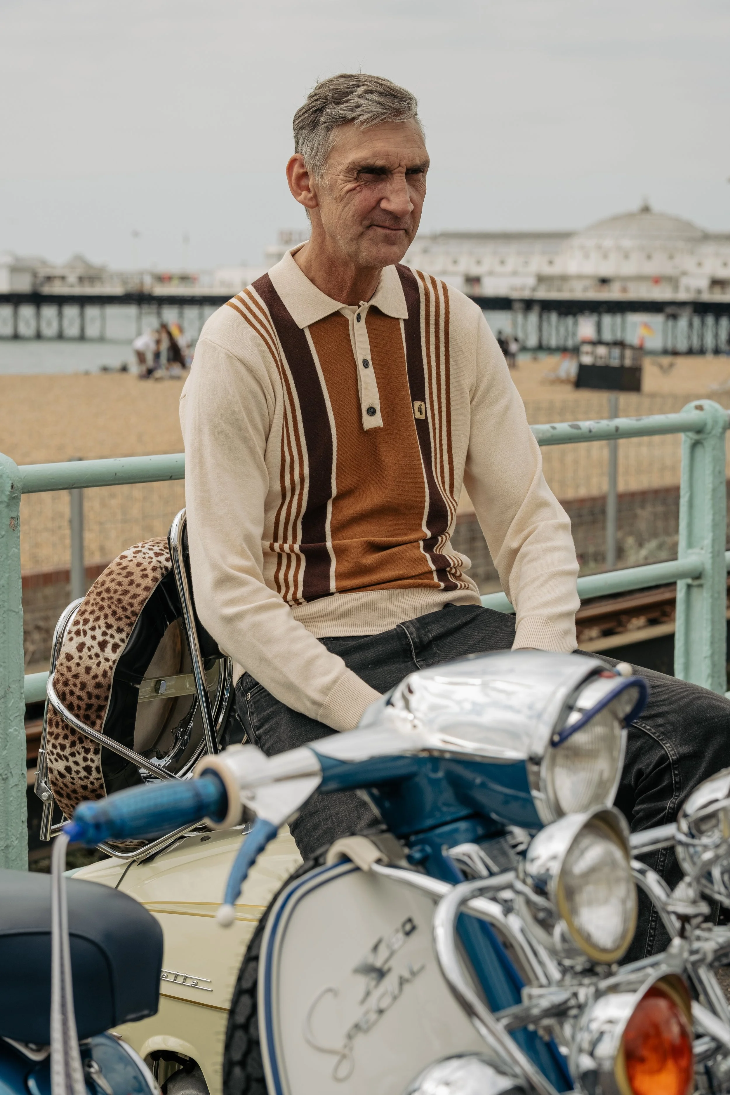 An older man sitting outdoors near a pier, with a vintage motorcycle and a leopard print bag, on a cloudy day.