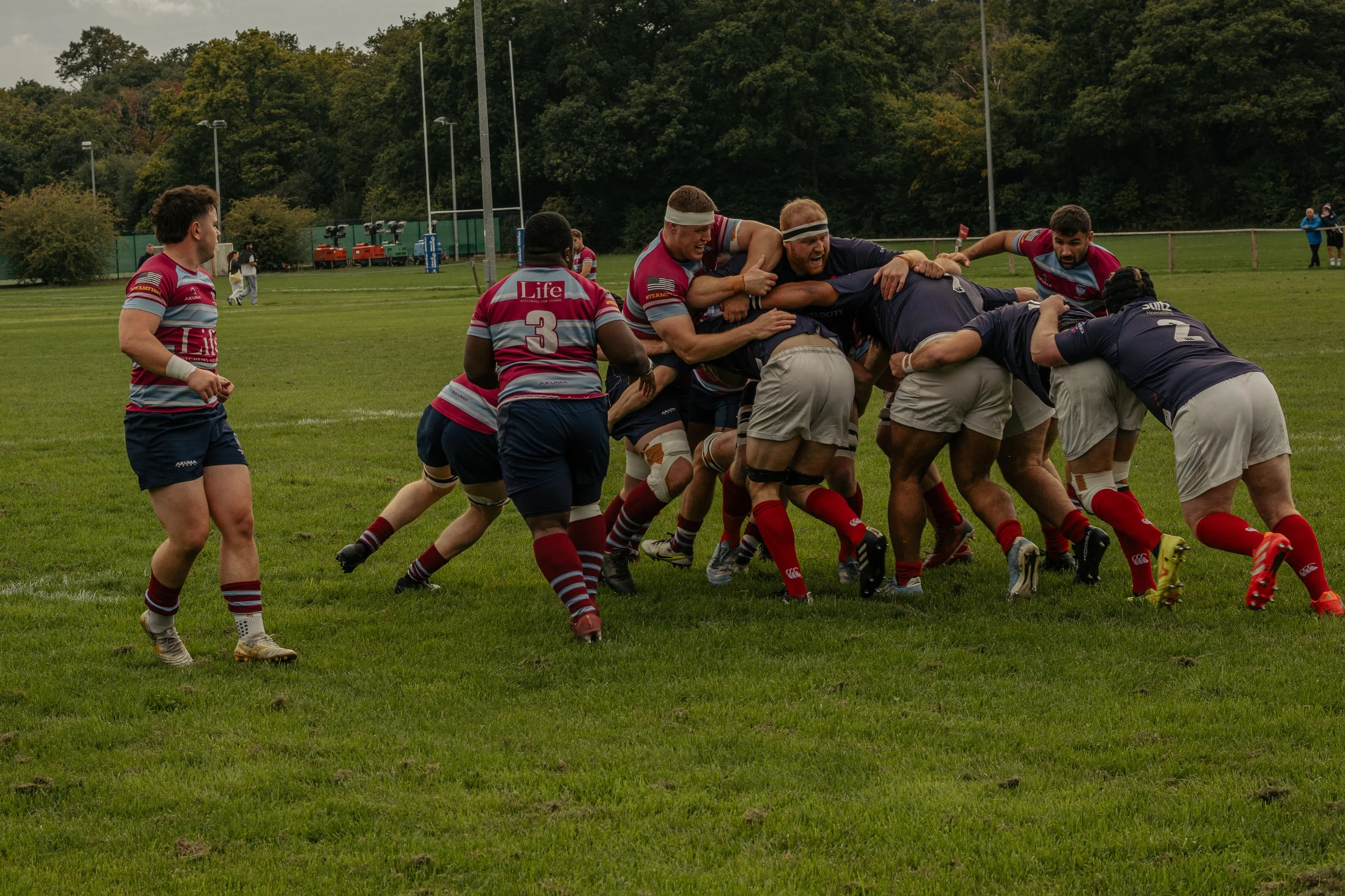 Rugby players engaged in a scrum on a grassy field during a game, with players wearing colorful jerseys and shorts.