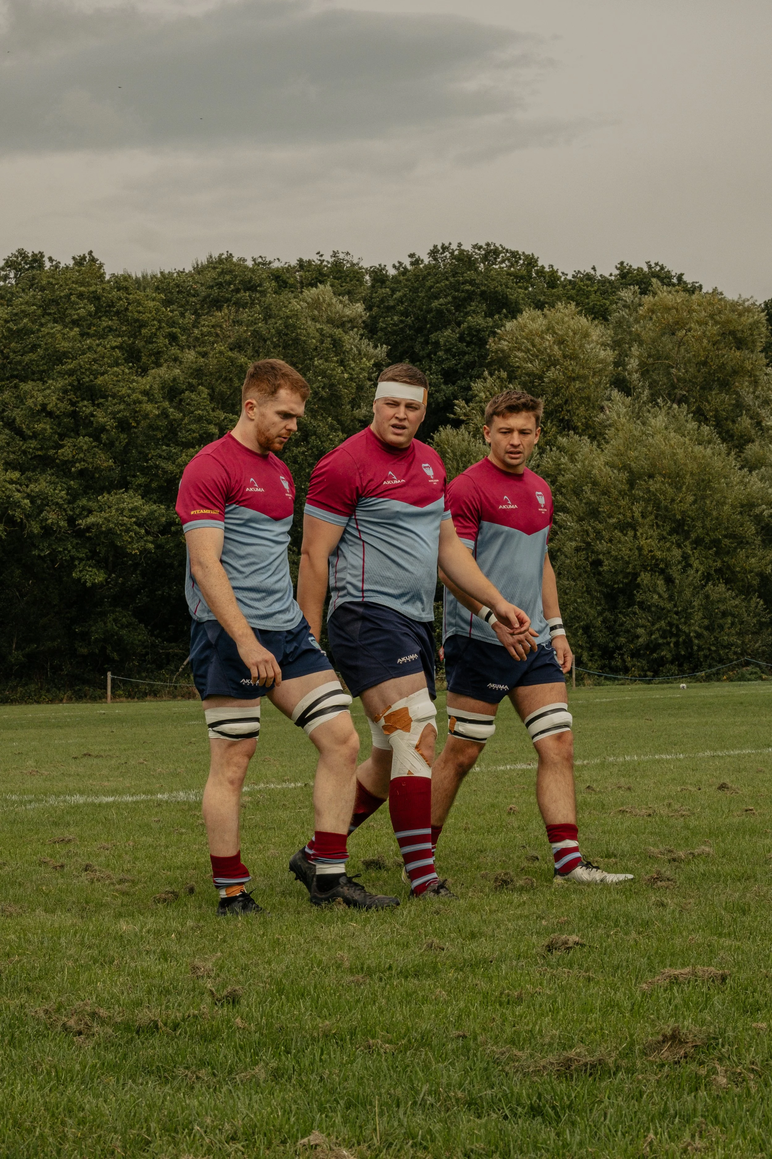 Three rugby players in maroon and blue jerseys walking on a grassy field, with trees and overcast sky in background.