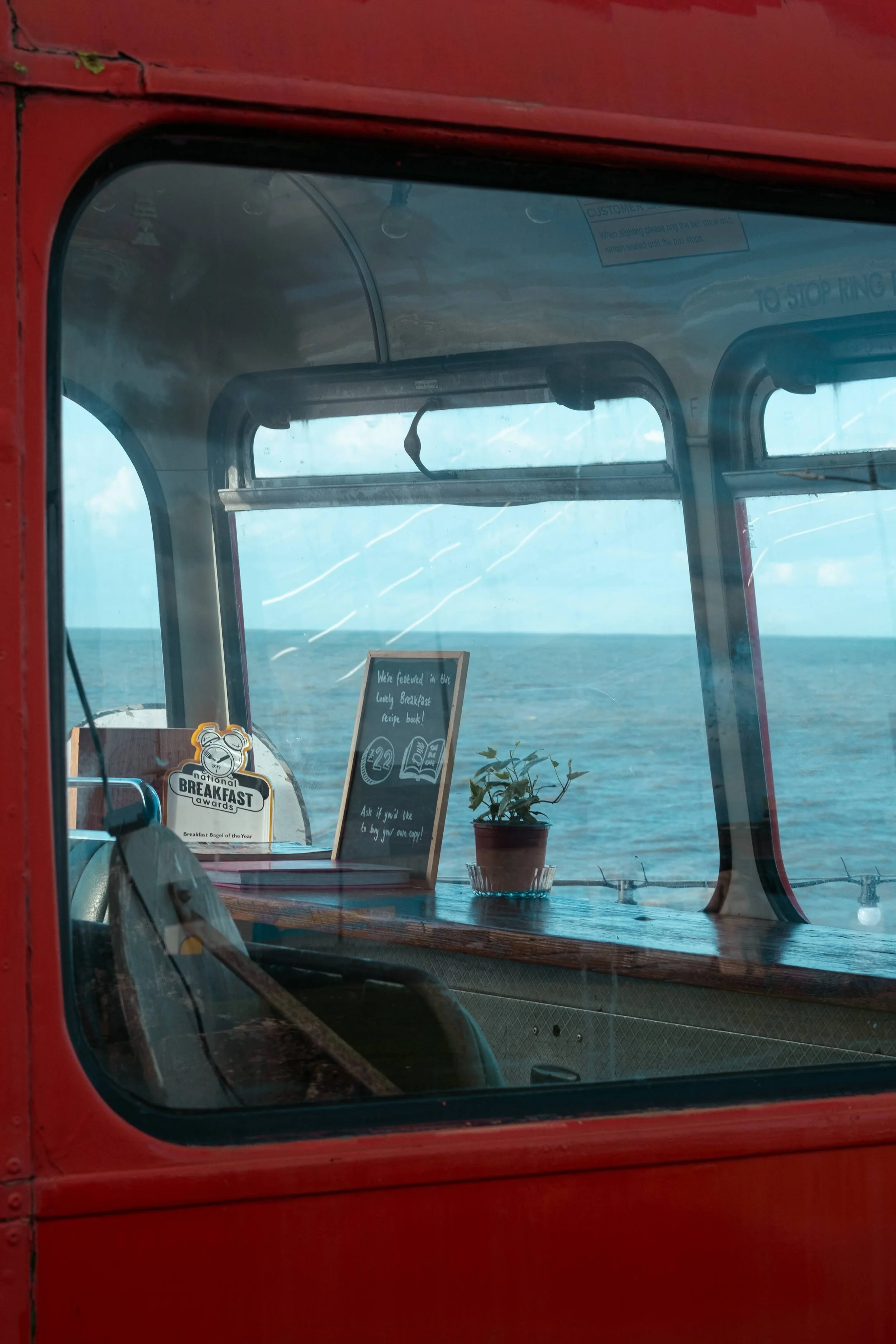 View through a window showing a small potted plant, a sign, and some awards on a wooden surface, with ocean and sky visible in the background.