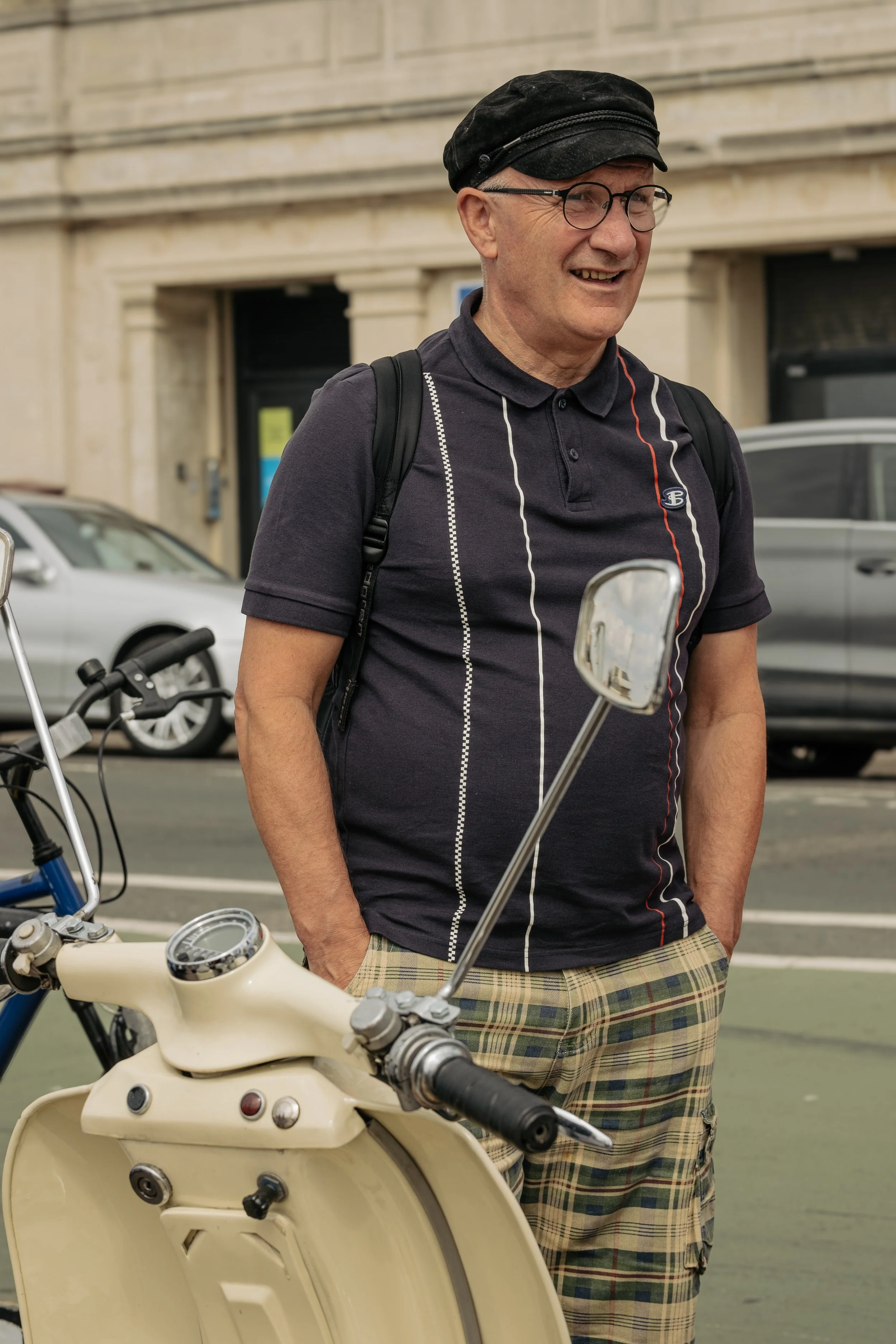 A man smiling with glasses and a black cap, standing next to a vintage cream-colored scooter on a city street.