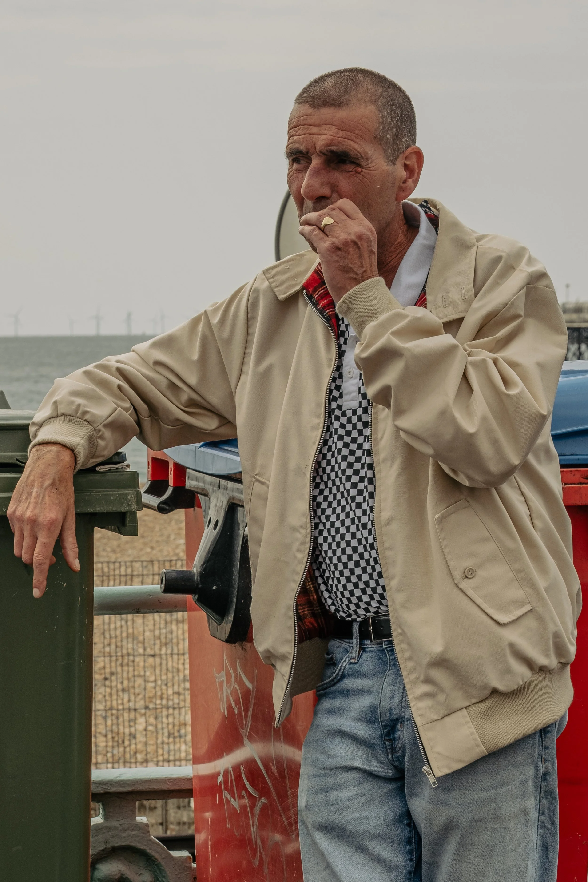 A middle-aged man with short hair wearing a beige jacket, checkered shirt, and jeans, standing outdoors near the coast, leaning on a green trash bin and holding his nose.
