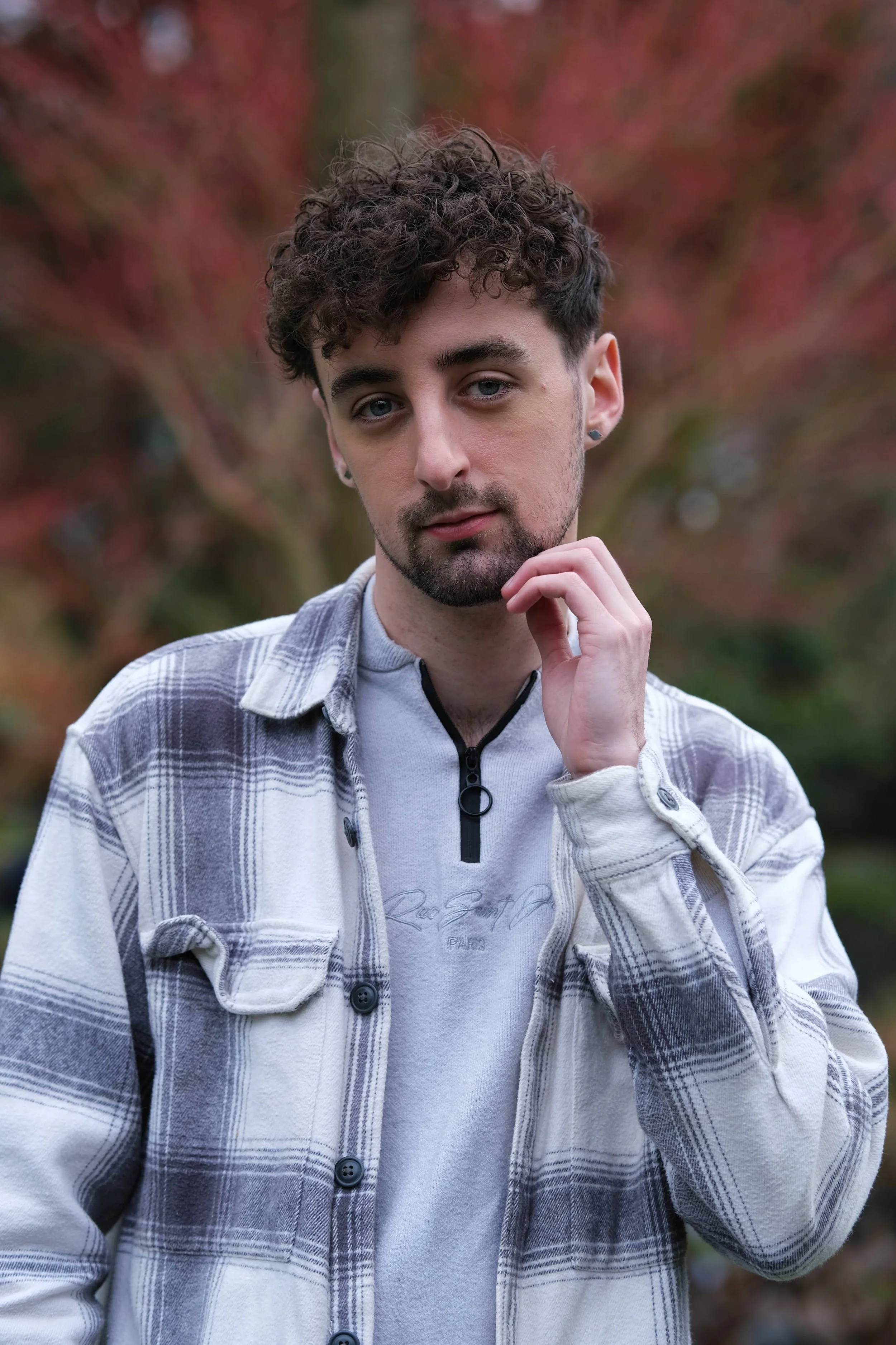 A young man with curly hair and light skin posing outdoors in front of blurry autumn-colored trees, wearing a gray and white checkered jacket over a gray top.