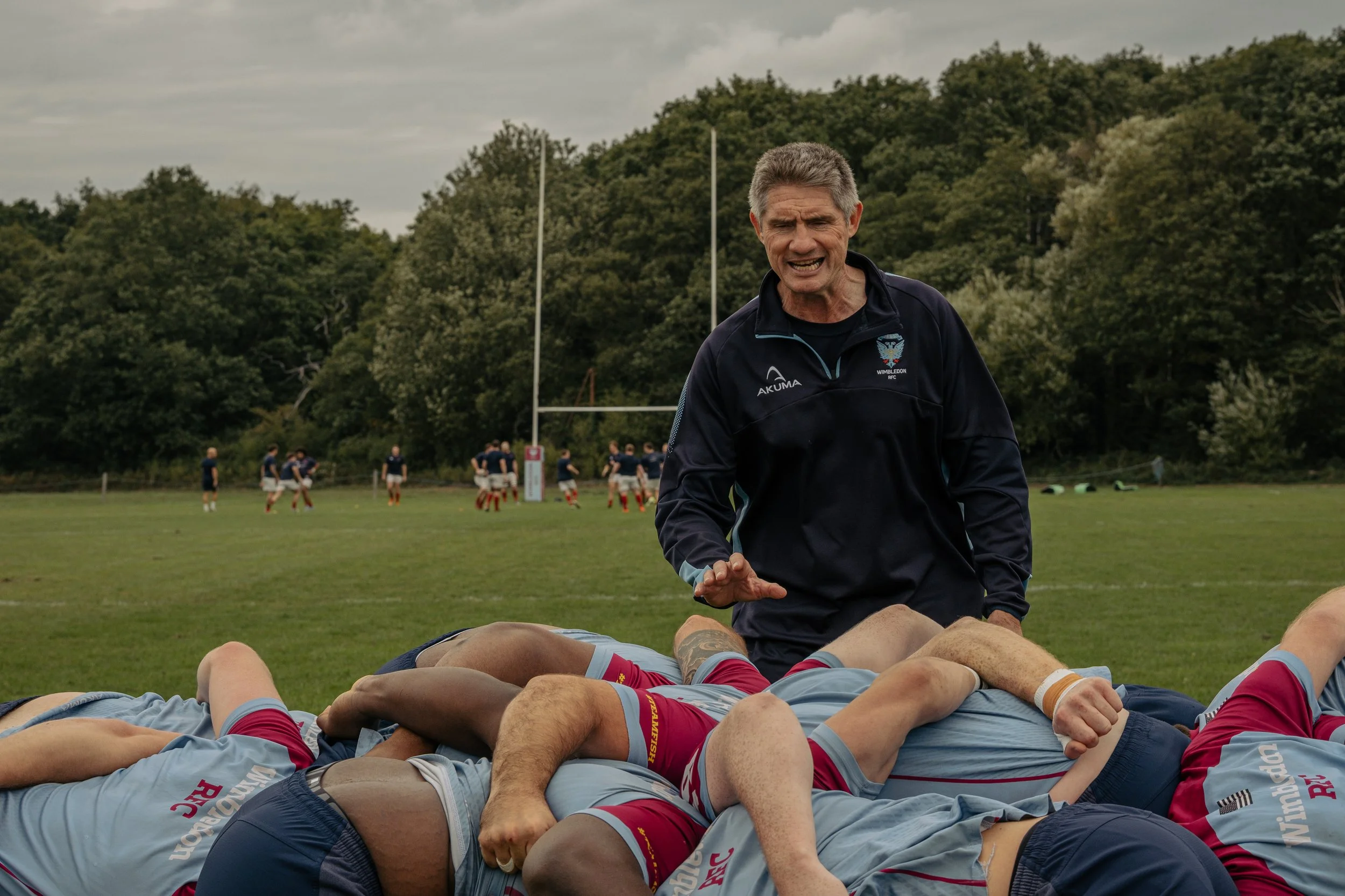 A rugby coach with gray hair and a dark sports jacket is giving instructions to players on the field during a match or practice, with a line of players in rugby uniforms in a ruck formation on the ground and other players in the background.