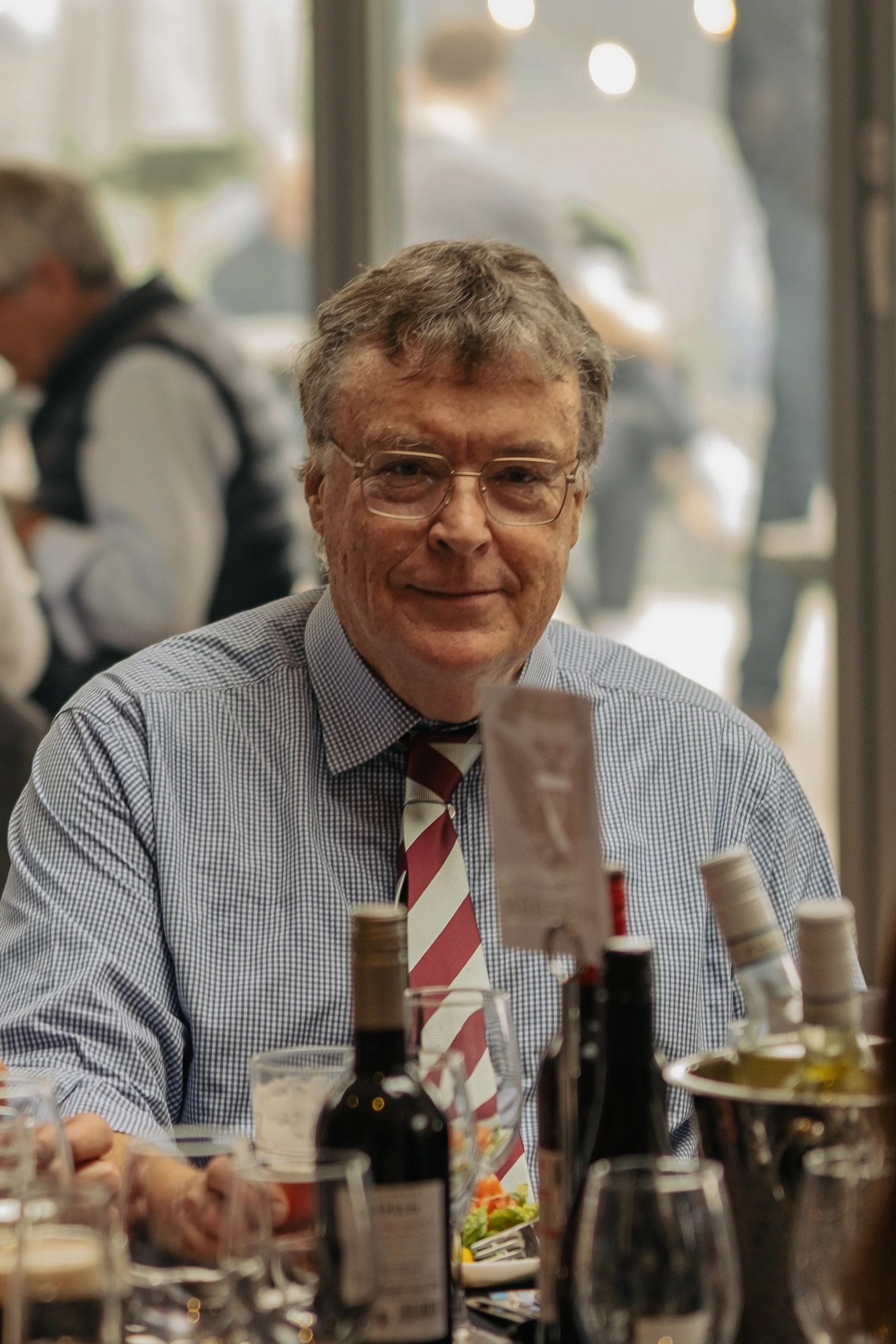 A man with glasses and a striped maroon and white tie sitting at a table with wine bottles and glasses in a restaurant or cafe, smiling at the camera.