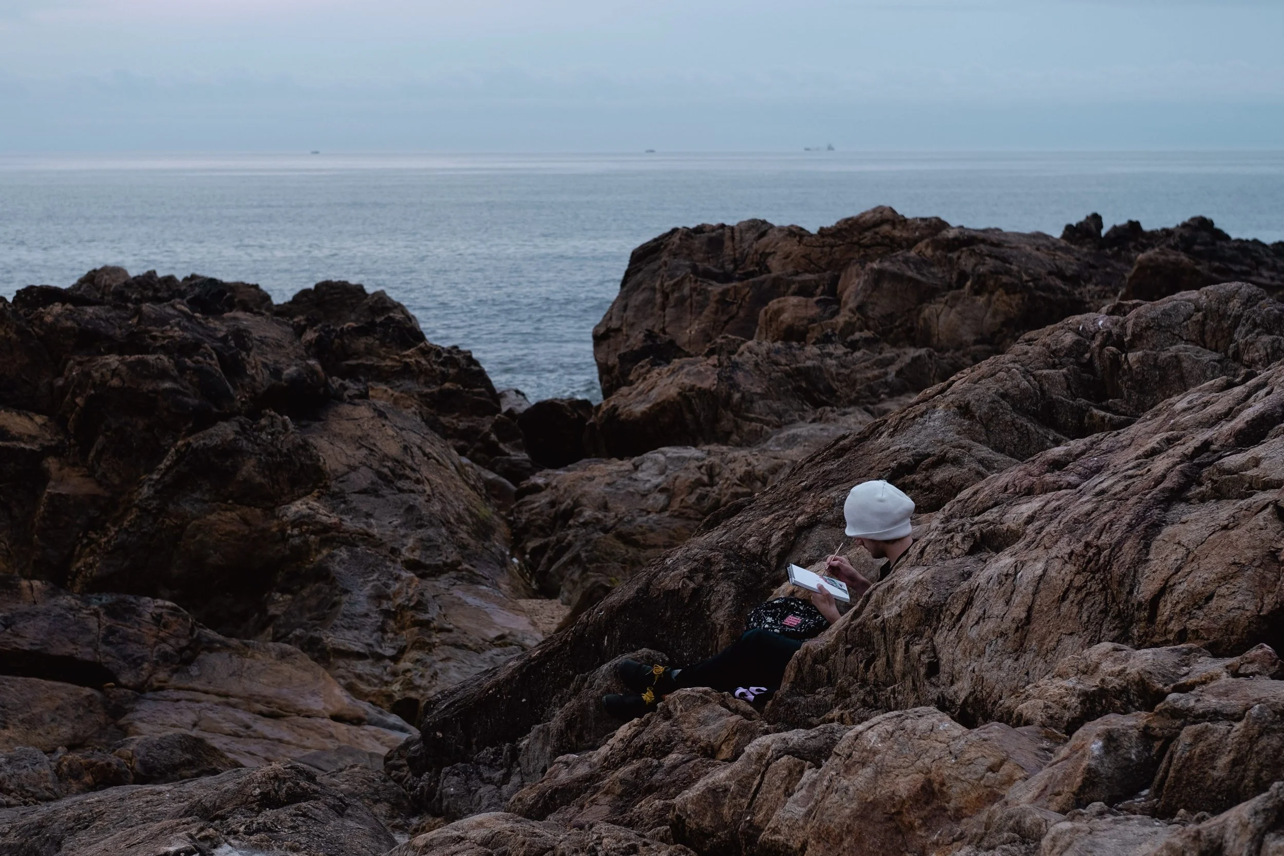 Person sitting on rocky shoreline, wearing white hat, reading or drawing in a notebook, with ocean in the background.