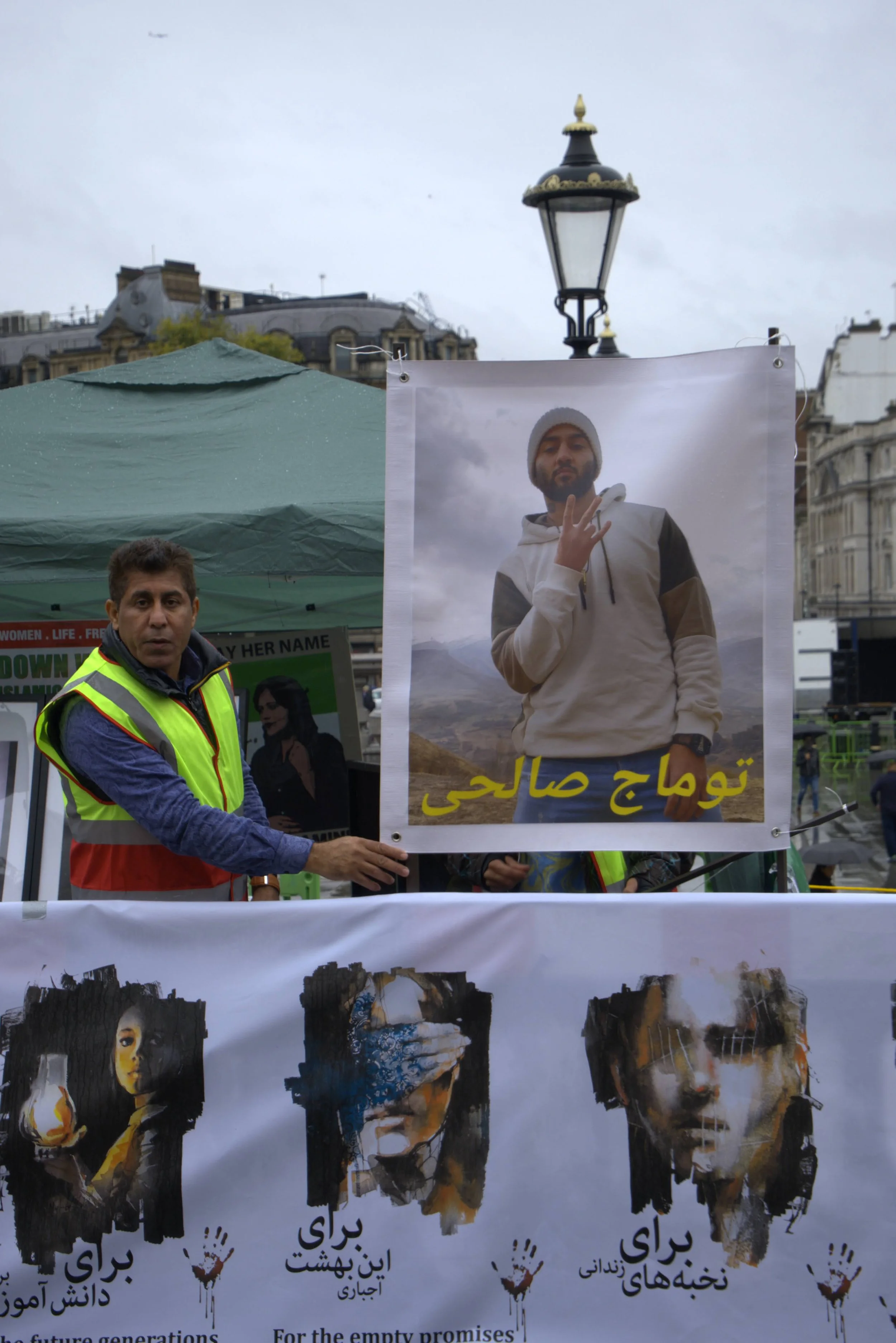 A man wearing a yellow safety vest pointing to a large poster of a young man with a beard, wearing a white hoodie and holding up a hand gesture, with Arabic text below. There is a green tent in the background, and the scene appears to be taking place