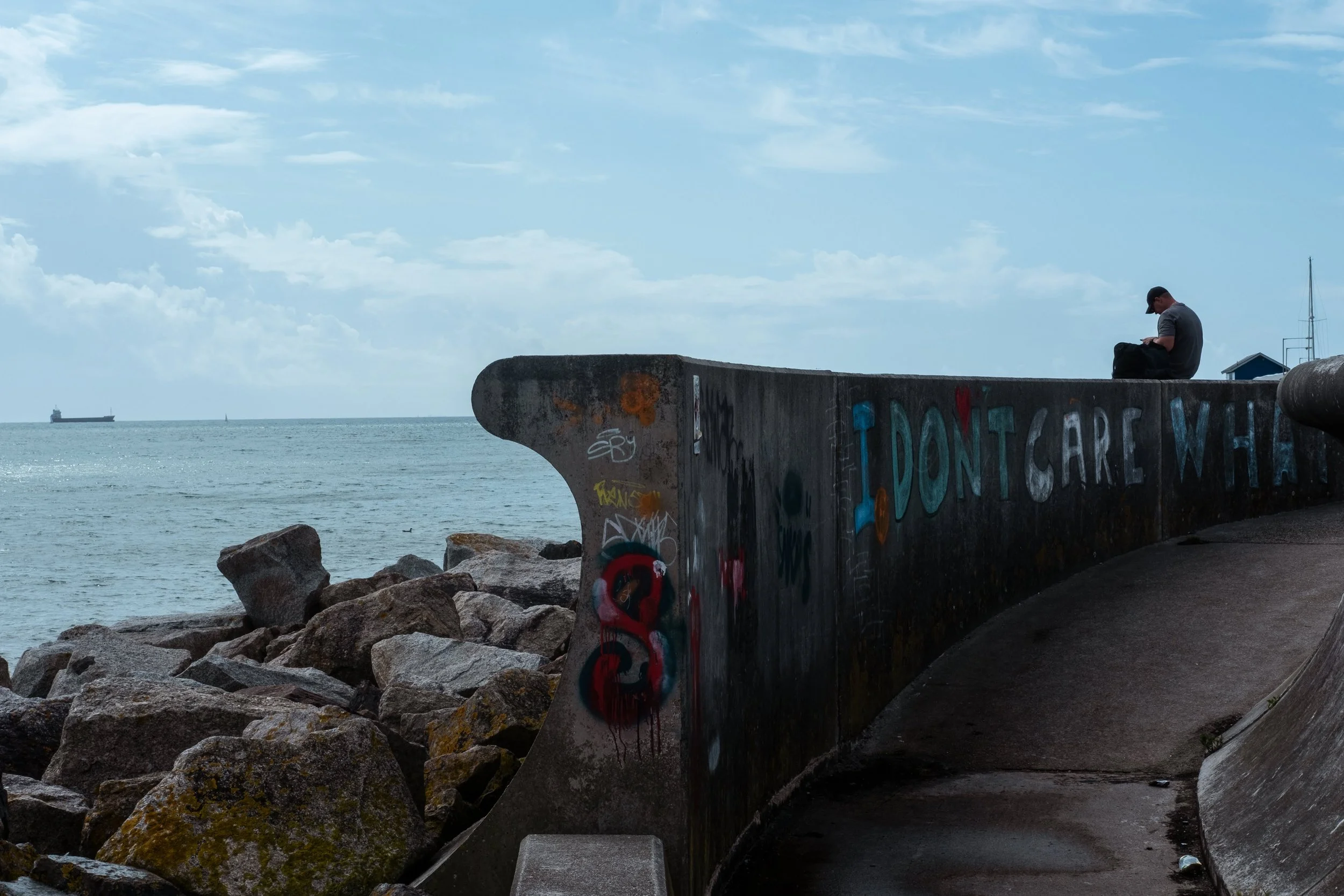 A man sitting on a graffiti-covered concrete barrier on a seaside pier, with large rocks and the ocean in the background, and a ship in the distance.