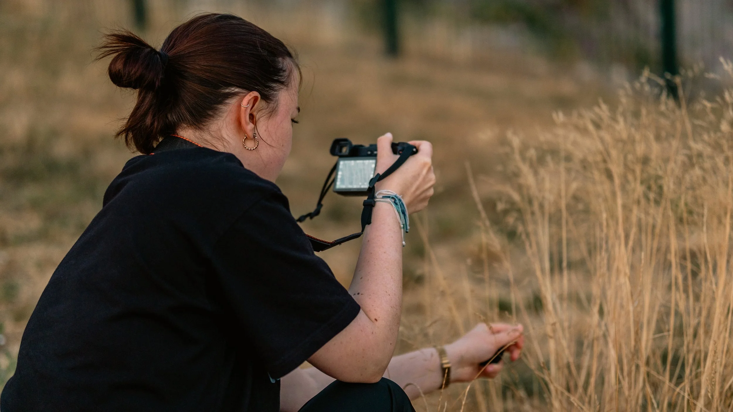 A woman with brown hair tied in a bun, wearing earrings and bracelets, is taking a photograph with a camera in a field of tall, dry grass.