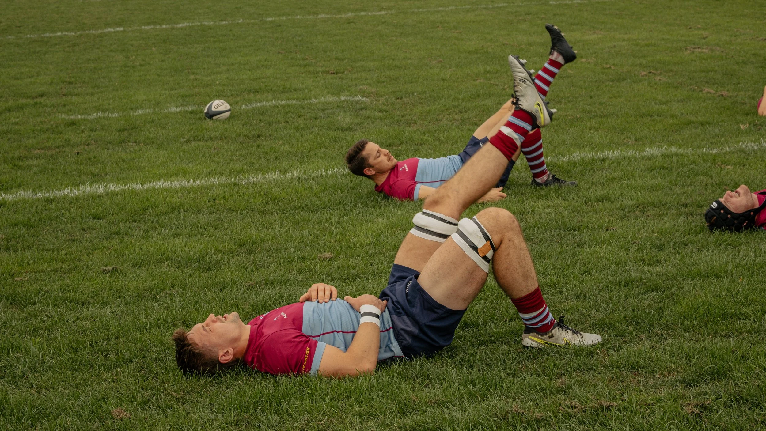 Two rugby players lying on the grass field, one on his back with knees bent, the other on his side, passing time during a break or after a match, with a rugby ball visible on the field.
