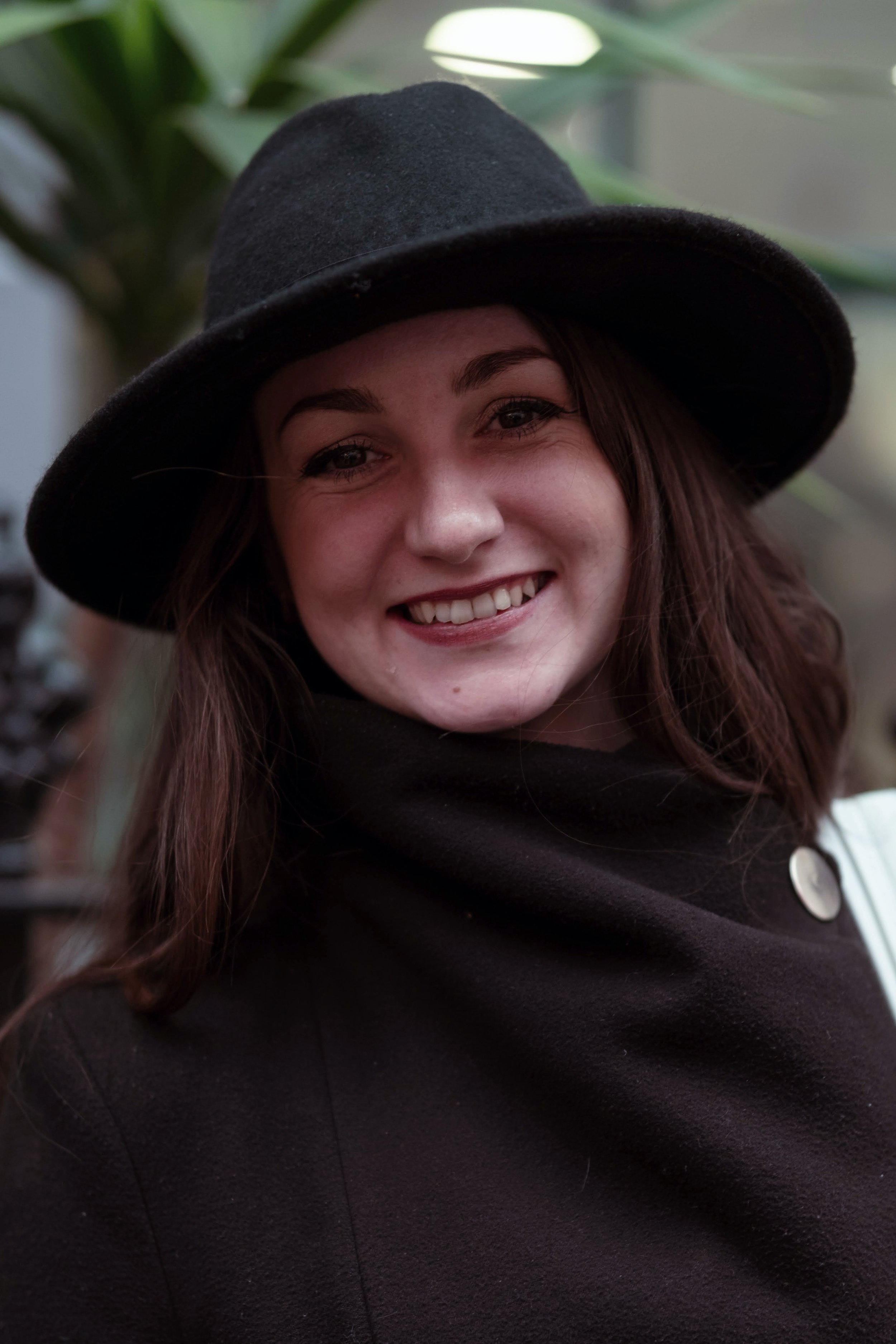A young woman smiling, wearing a black wide-brimmed hat and a black coat with a large button.