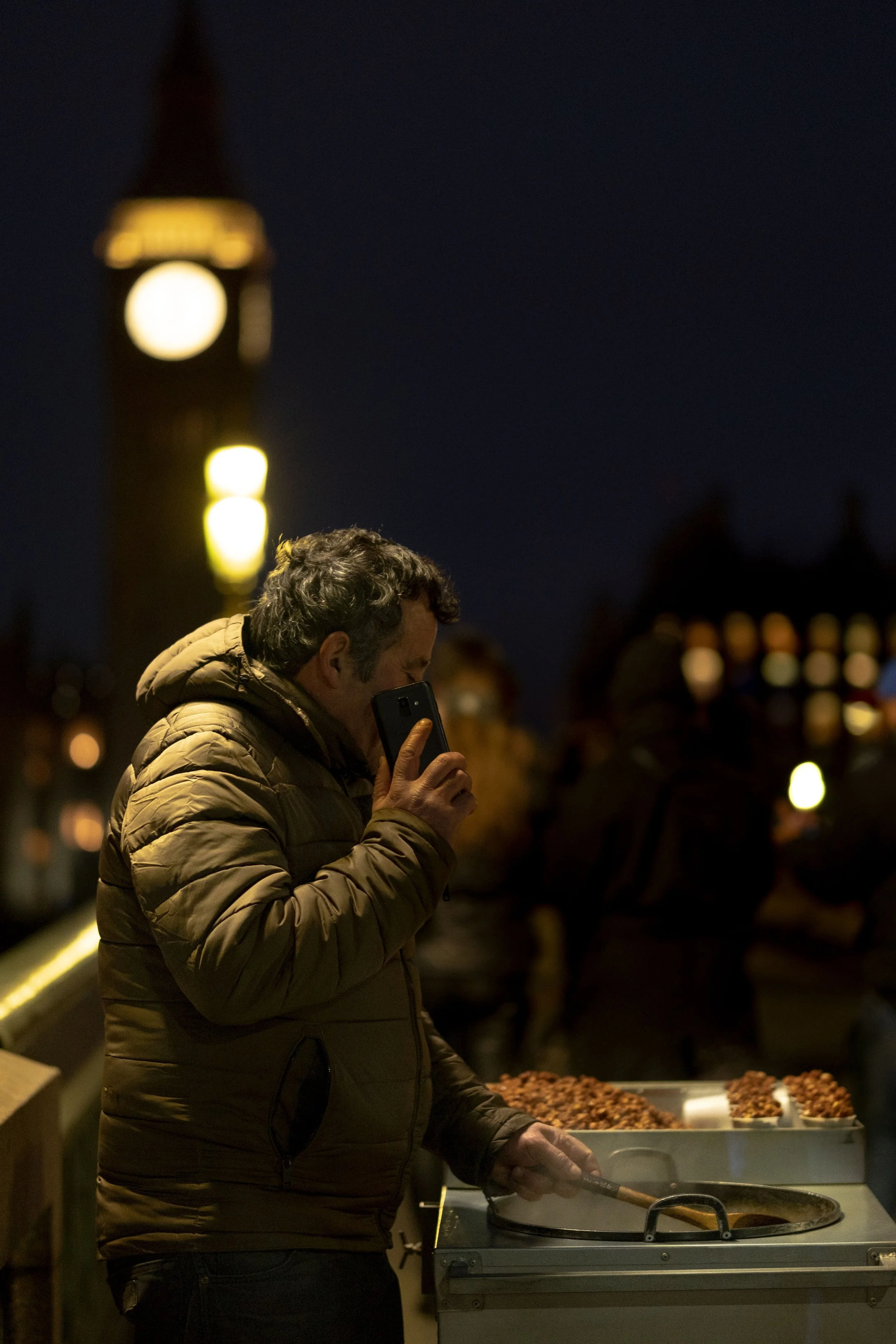 A man in a brown jacket cooking food at a street stall at night, with Big Ben in the background.