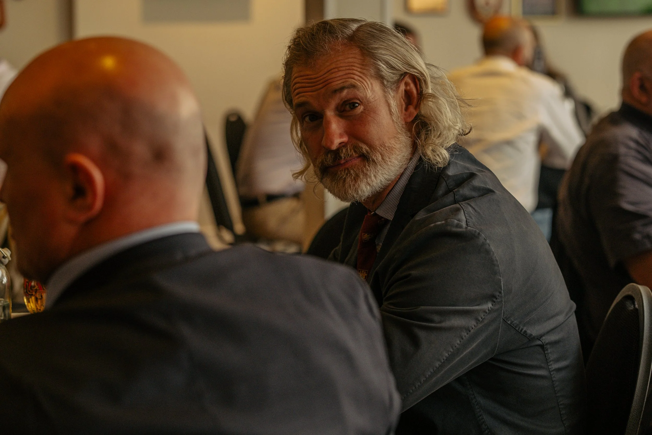 A man with gray hair and a beard is looking at the camera while sitting at a table during a gathering.