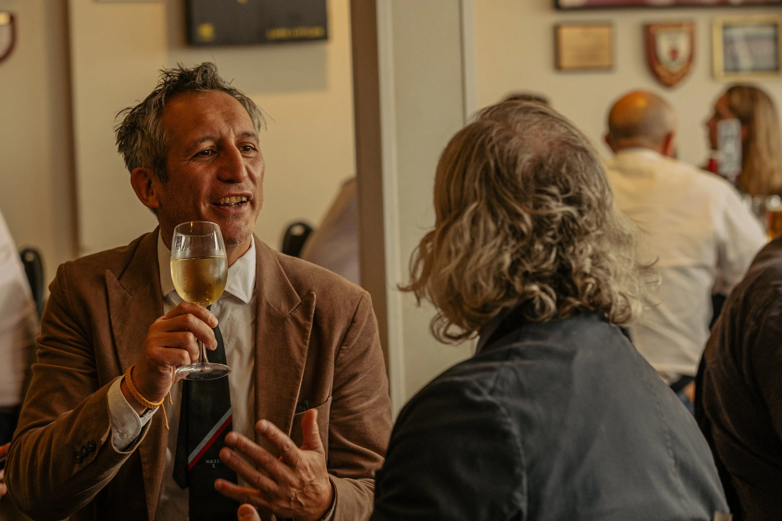 Man with glasses and gray hair holding a glass of white wine, engaging in conversation at a social gathering in a bar or restaurant, with other people and framed pictures on the wall in the background.