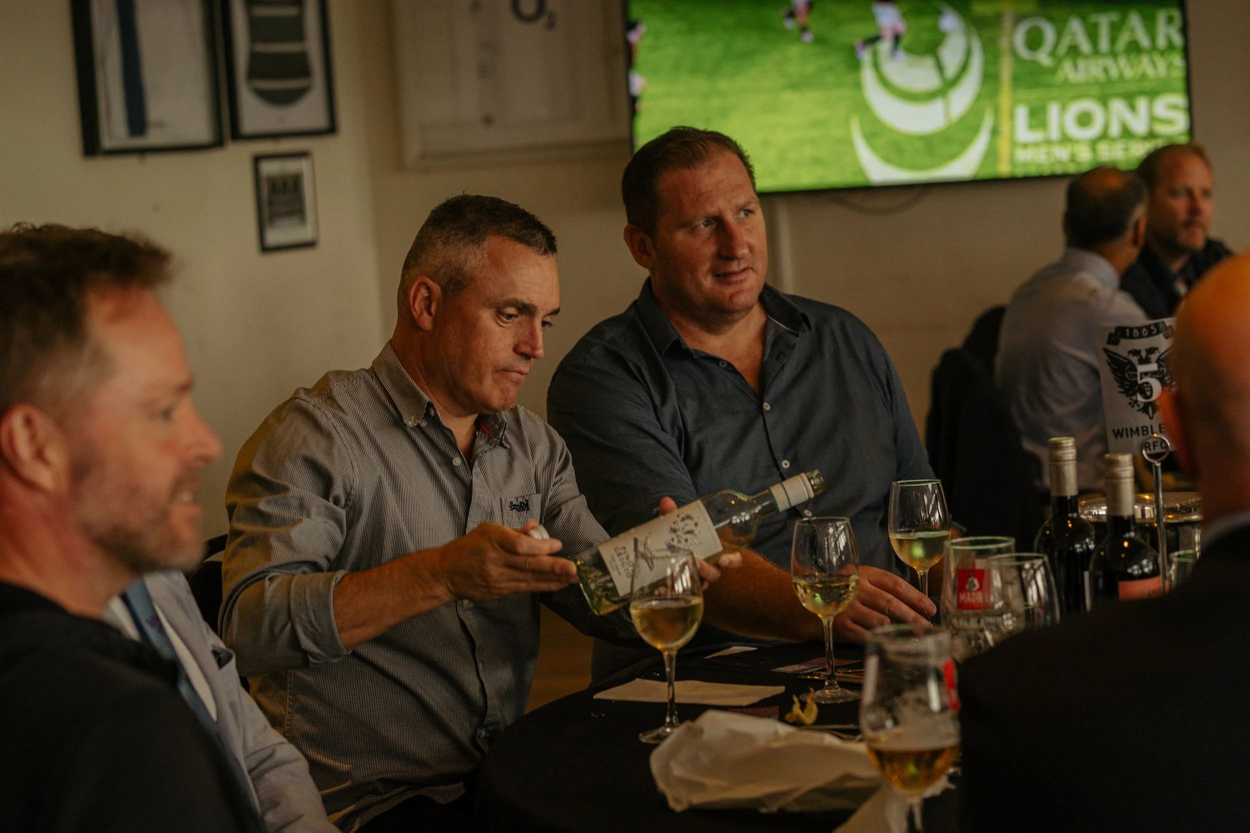 Men sitting at a table with wine glasses, with a large screen displaying the Qatar Airways Lions logo in the background.