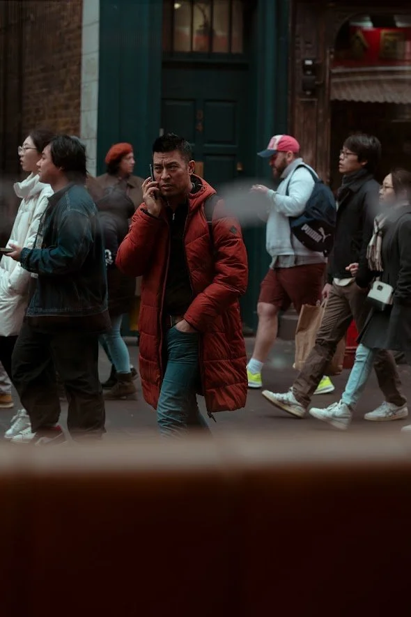 A man wearing a red jacket talking on his cellphone while walking through a busy street with many pedestrians.