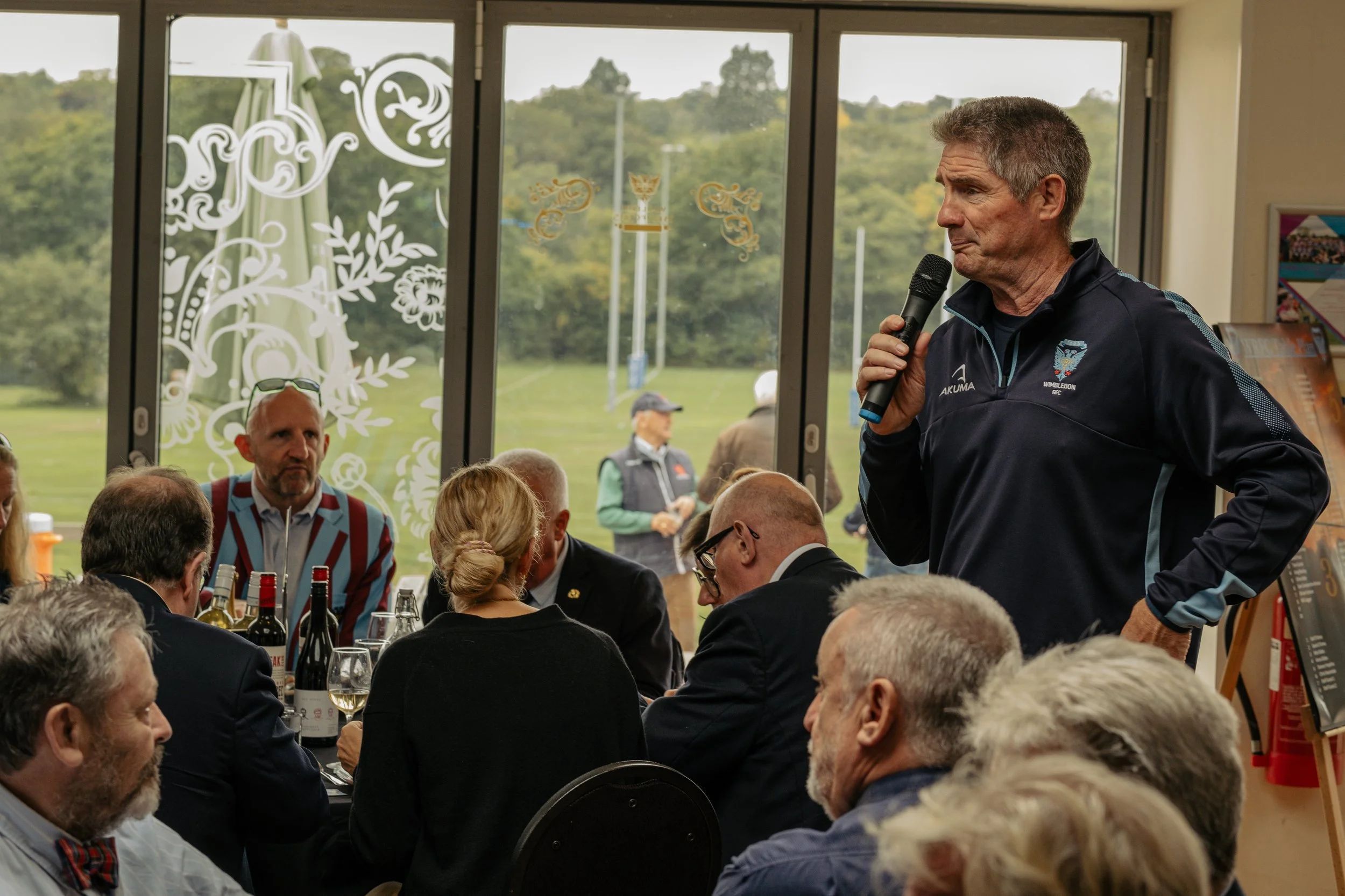 A man speaking into a microphone to a group of people seated around a table during an indoor event, with large windows showing an outdoor grassy area.