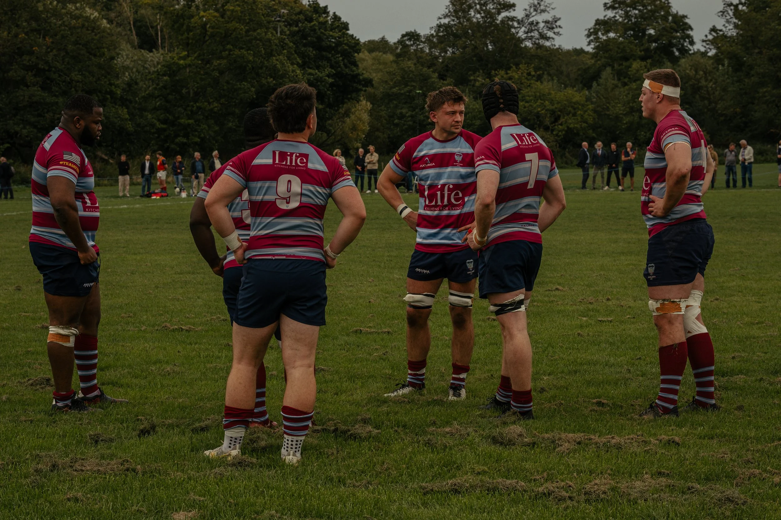 Rugby players in maroon and blue uniforms standing in a huddle on a grassy field, with spectators in the background.