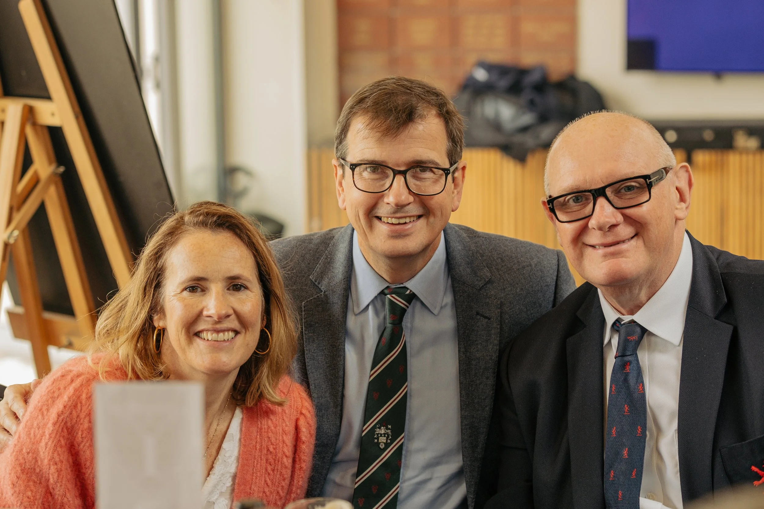 A group of three people, two men and one woman, smiling and posing closely together at a social gathering or event indoors.