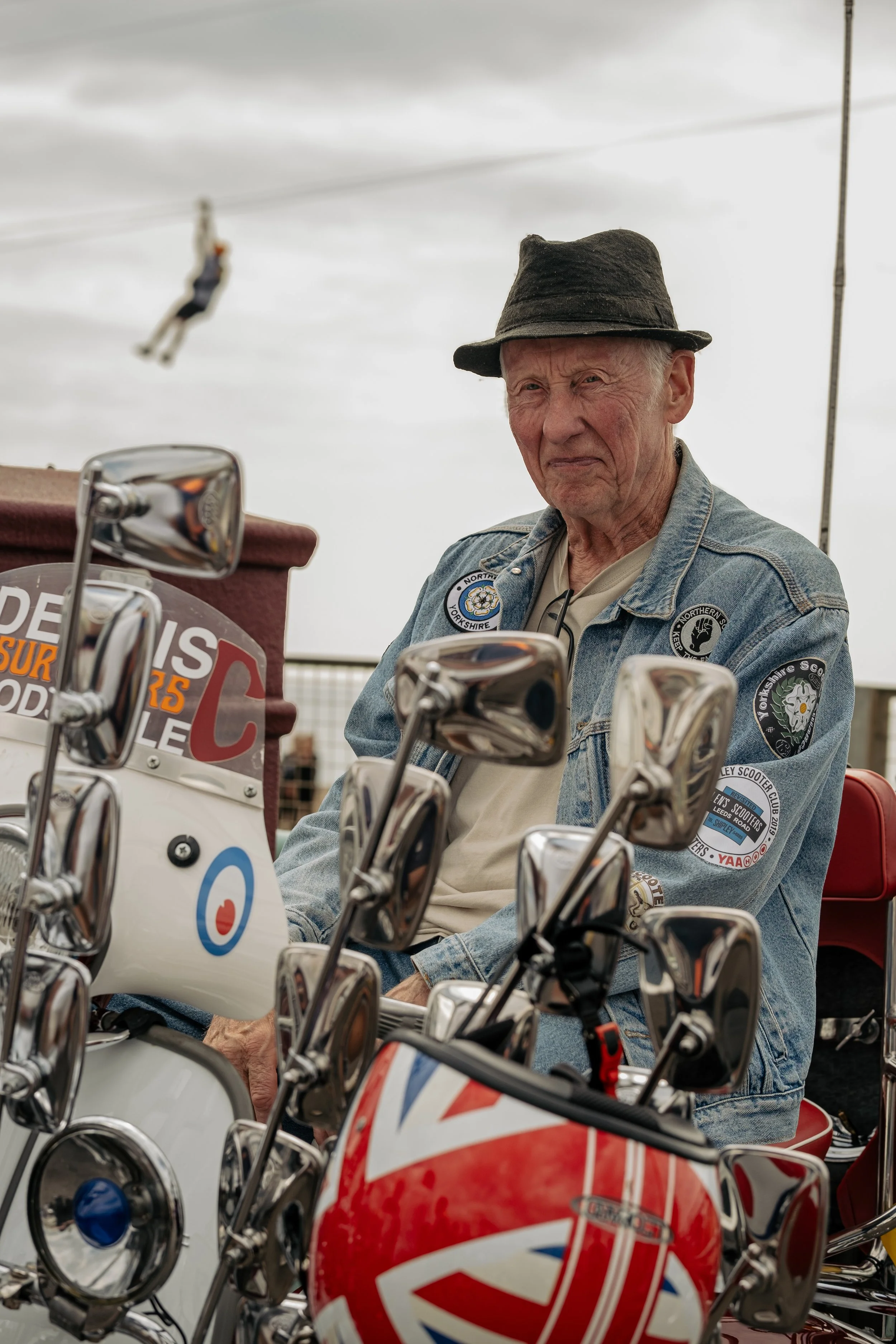 An elderly man wearing a black hat and denim jacket with patches, sitting on a scooter, with a cloudy sky and a person zip lining in the background.