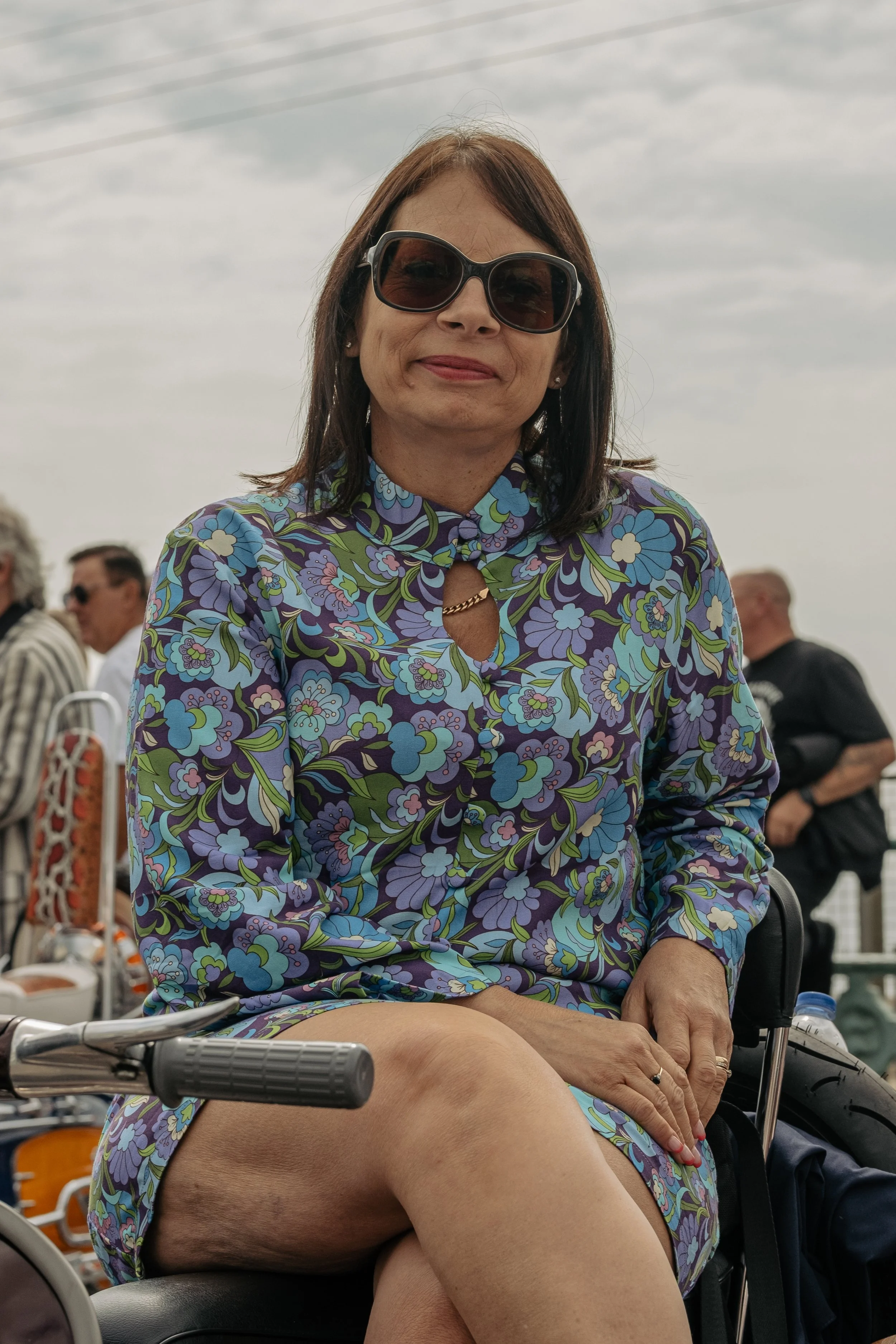Woman sitting outdoors wearing a floral dress and sunglasses, with people and bicycles in the background.