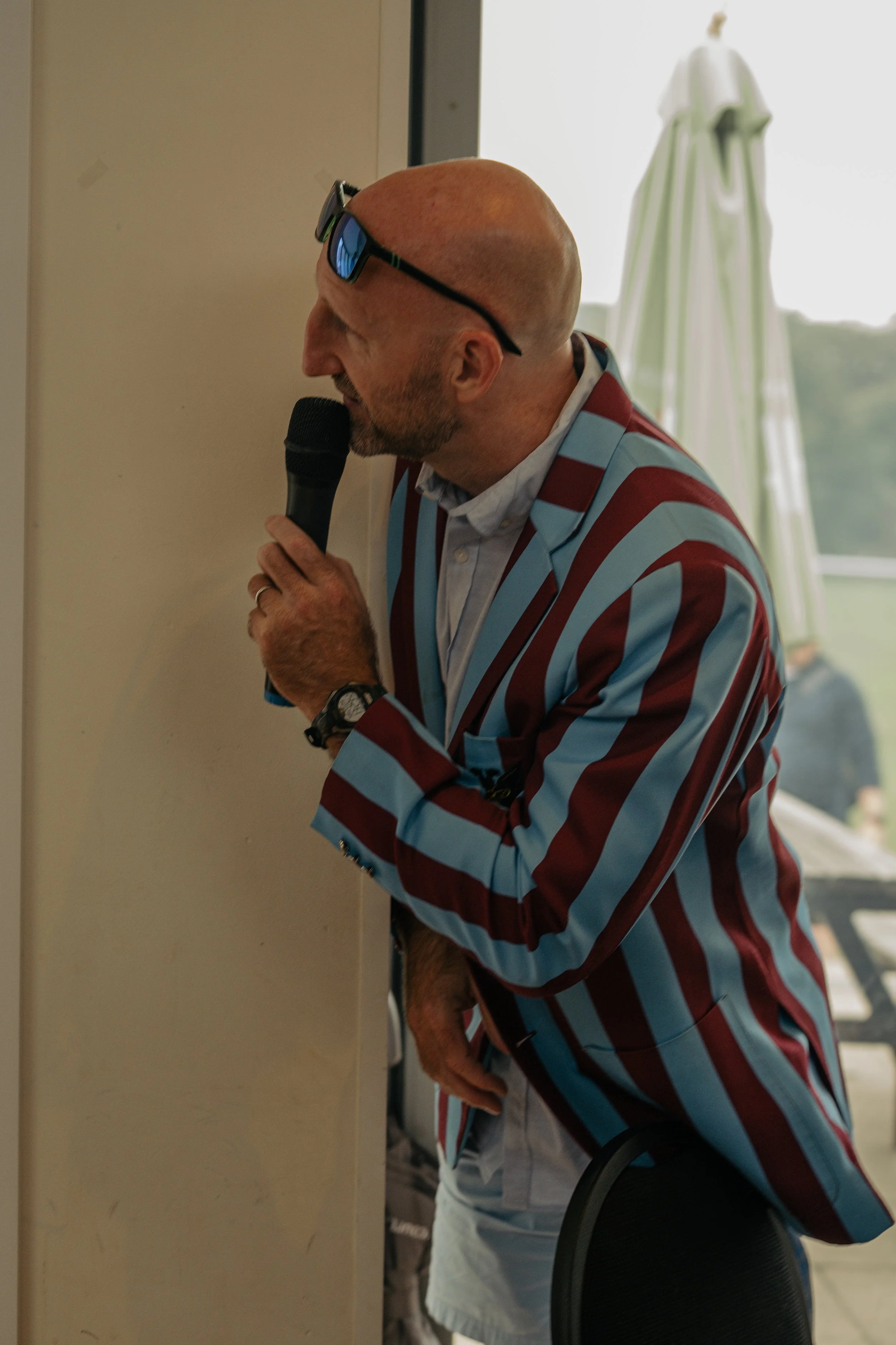 A man with a bald head and sunglasses resting on his head, wearing a jacket with red and blue vertical stripes, speaking into a microphone indoors near a window, with an outdoor patio and umbrella visible outside.