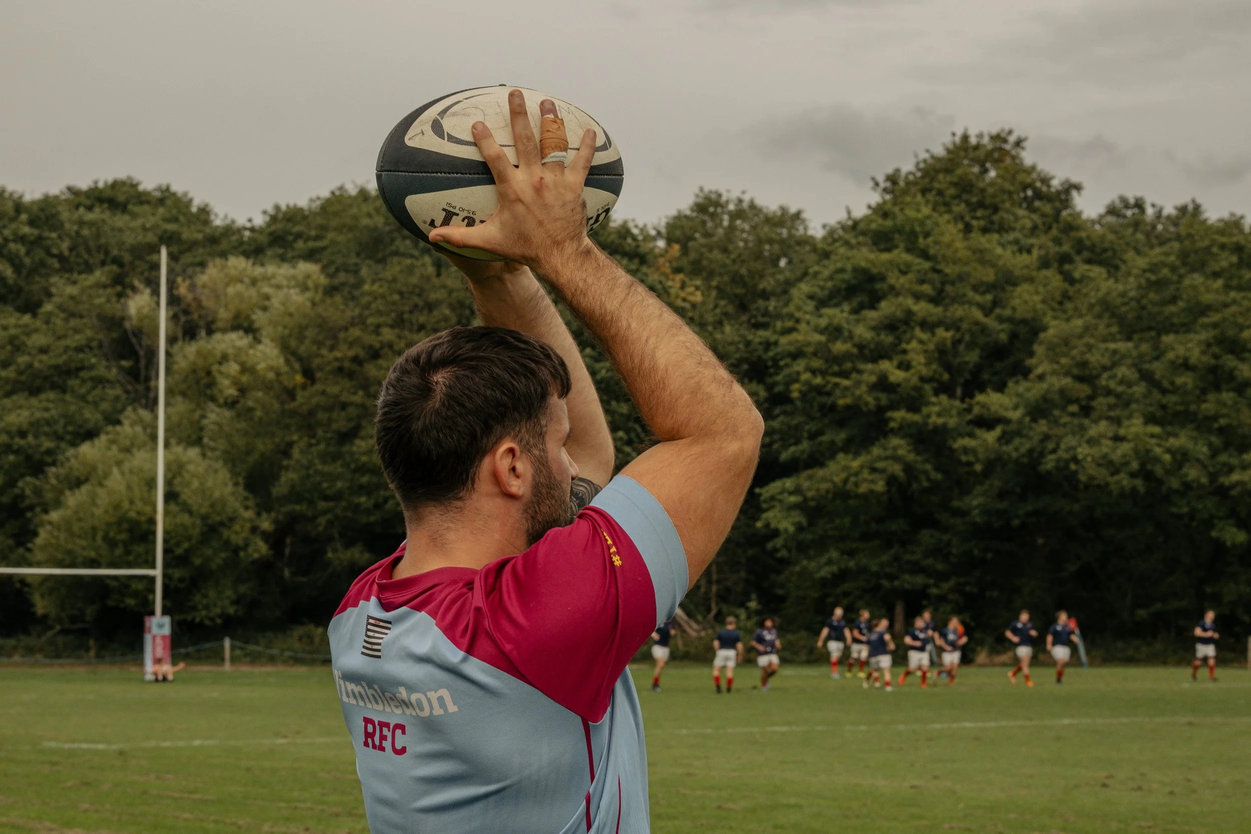 A rugby player is preparing to kick the ball, with a team in the background on a green field and trees surrounding the field.