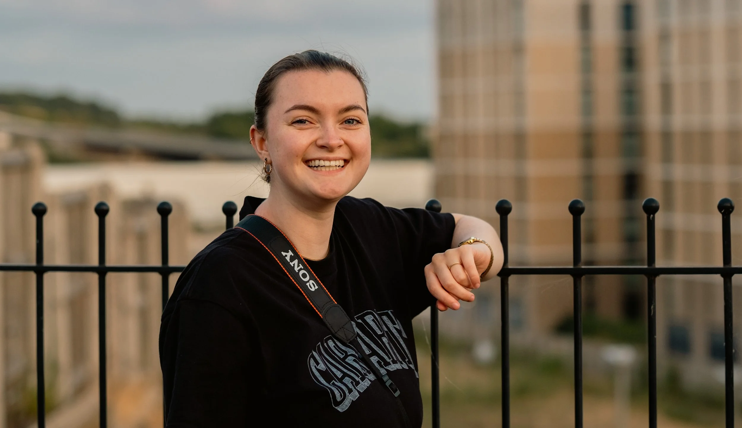 A young woman with dark hair tied back, smiling, wearing a black T-shirt with white lettering, standing outdoors behind a black metal fence, with city buildings and a cloudy sky in the background.