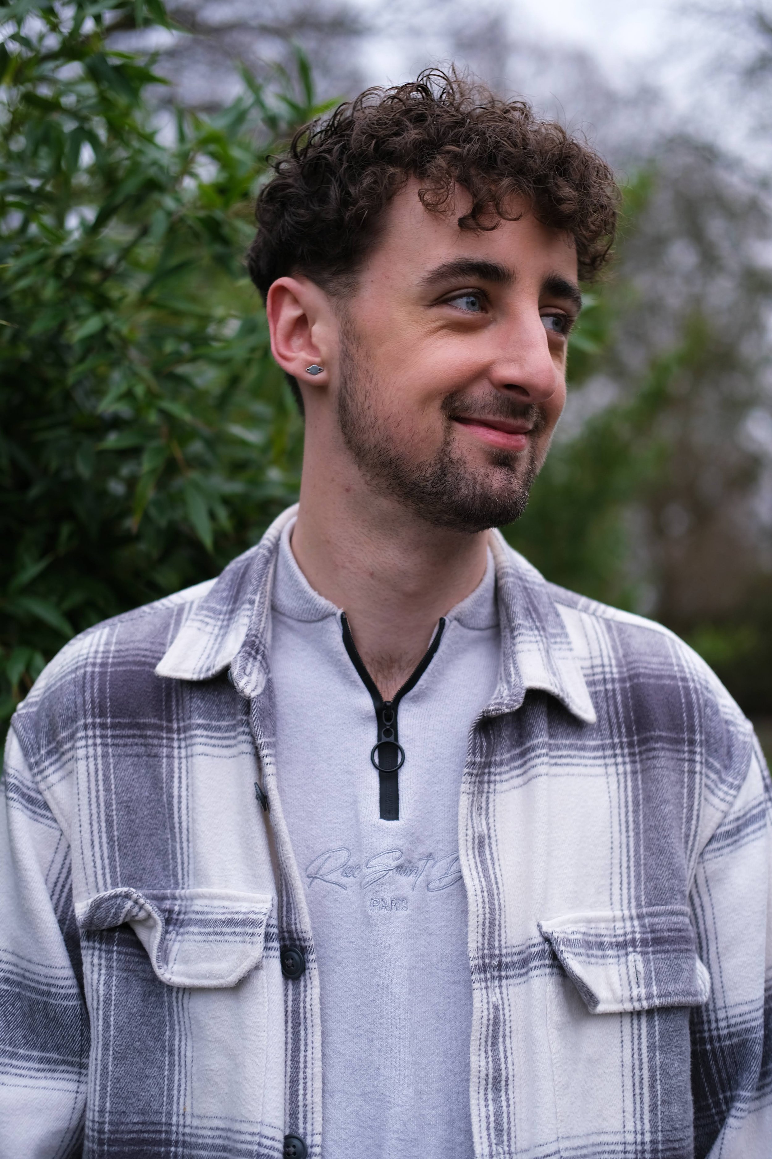 A young man with curly hair, wearing a light gray quarter-zip shirt and a plaid jacket, standing outdoors with green foliage in the background.