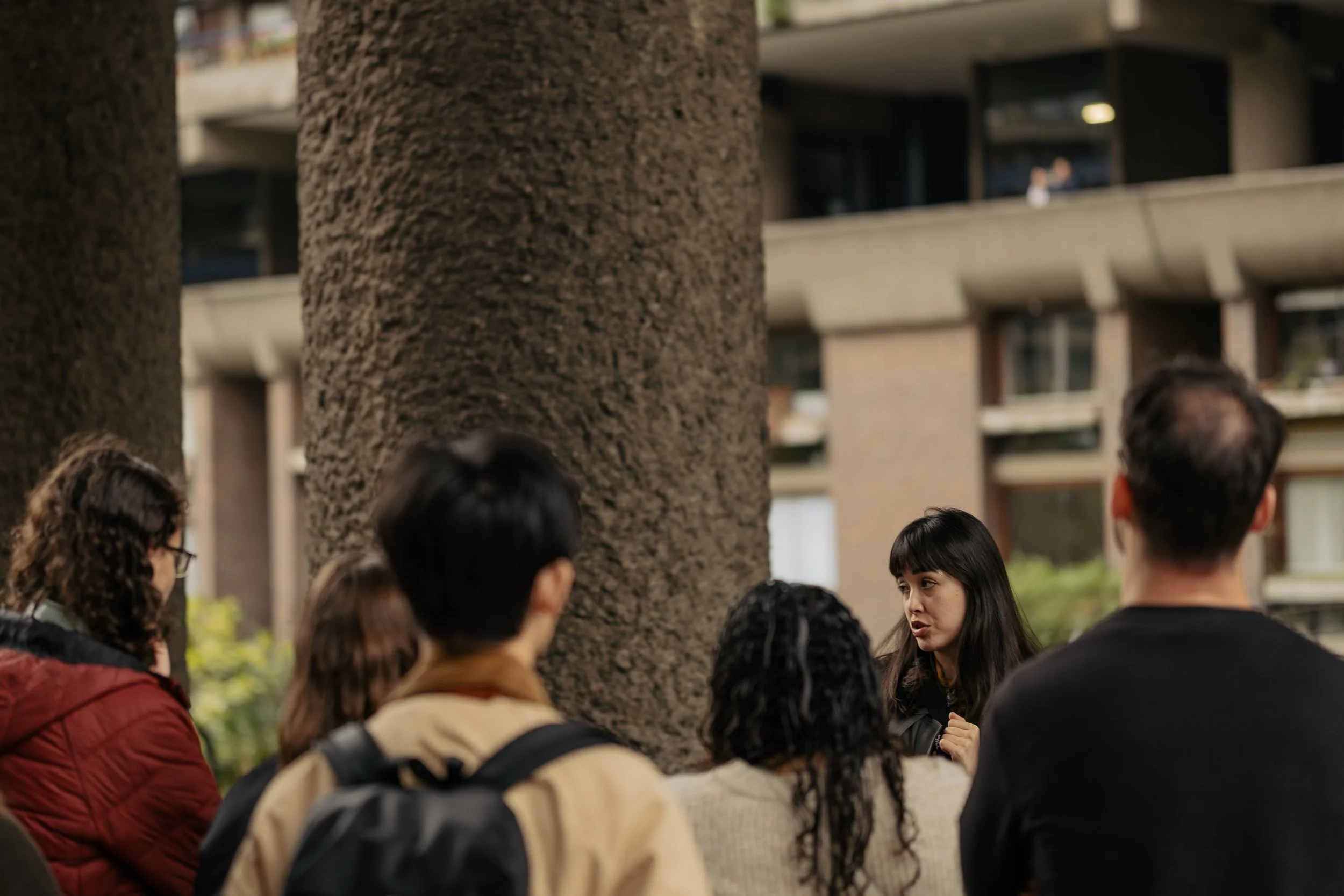 A group of people gathered outdoors around a large tree trunk, with a woman speaking to them and a modern building in the background.