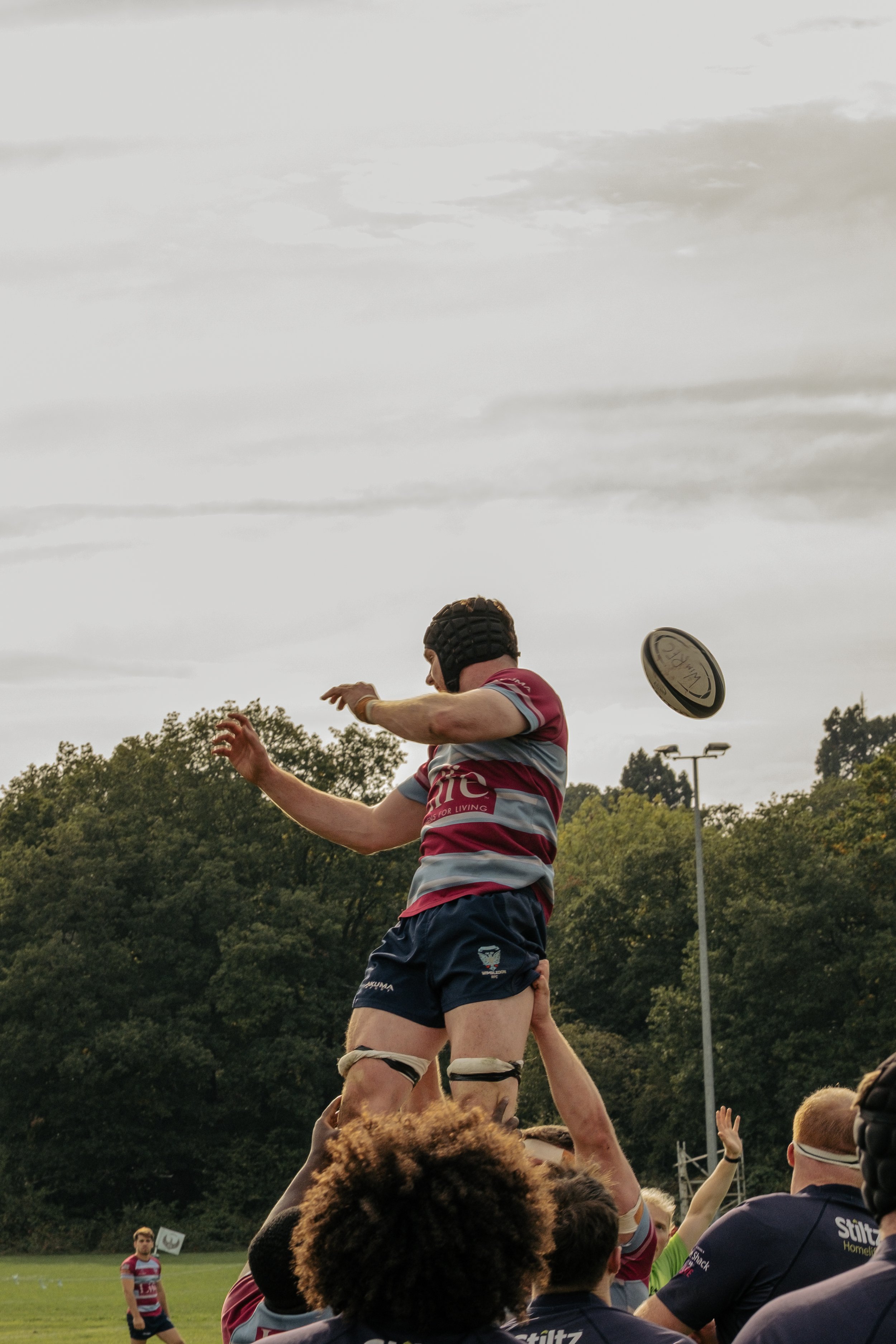 Rugby player in maroon and light blue jersey catches a rugby ball during a lineout in a game, with teammates and a referee on a grassy field and a cloudy sky overhead.