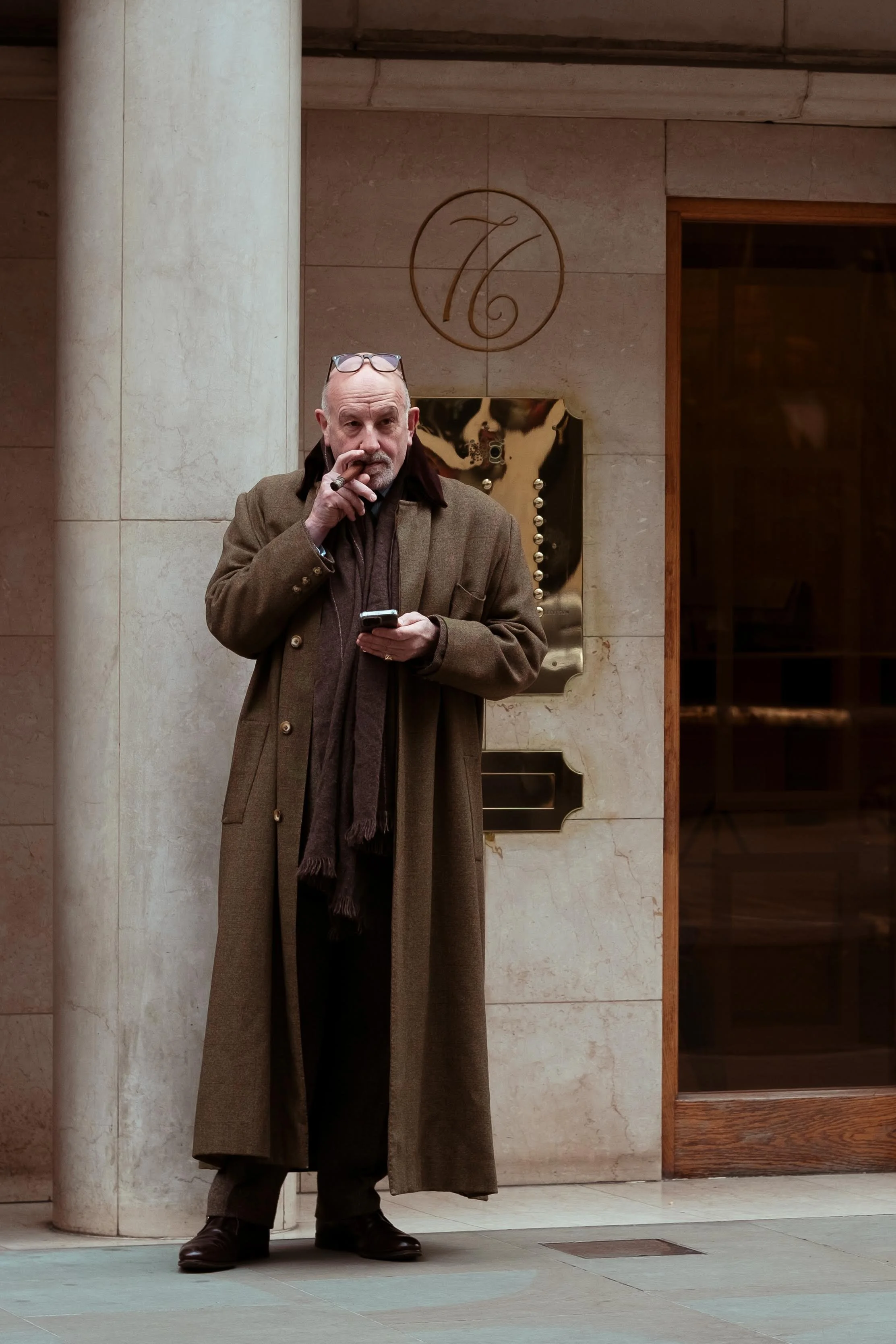 A middle-aged man with glasses on his head, wearing a long brown coat, standing outside in front of a beige marble wall with a circular decorative emblem. He is holding a smartphone in one hand and smoking a cigarette with the other.
