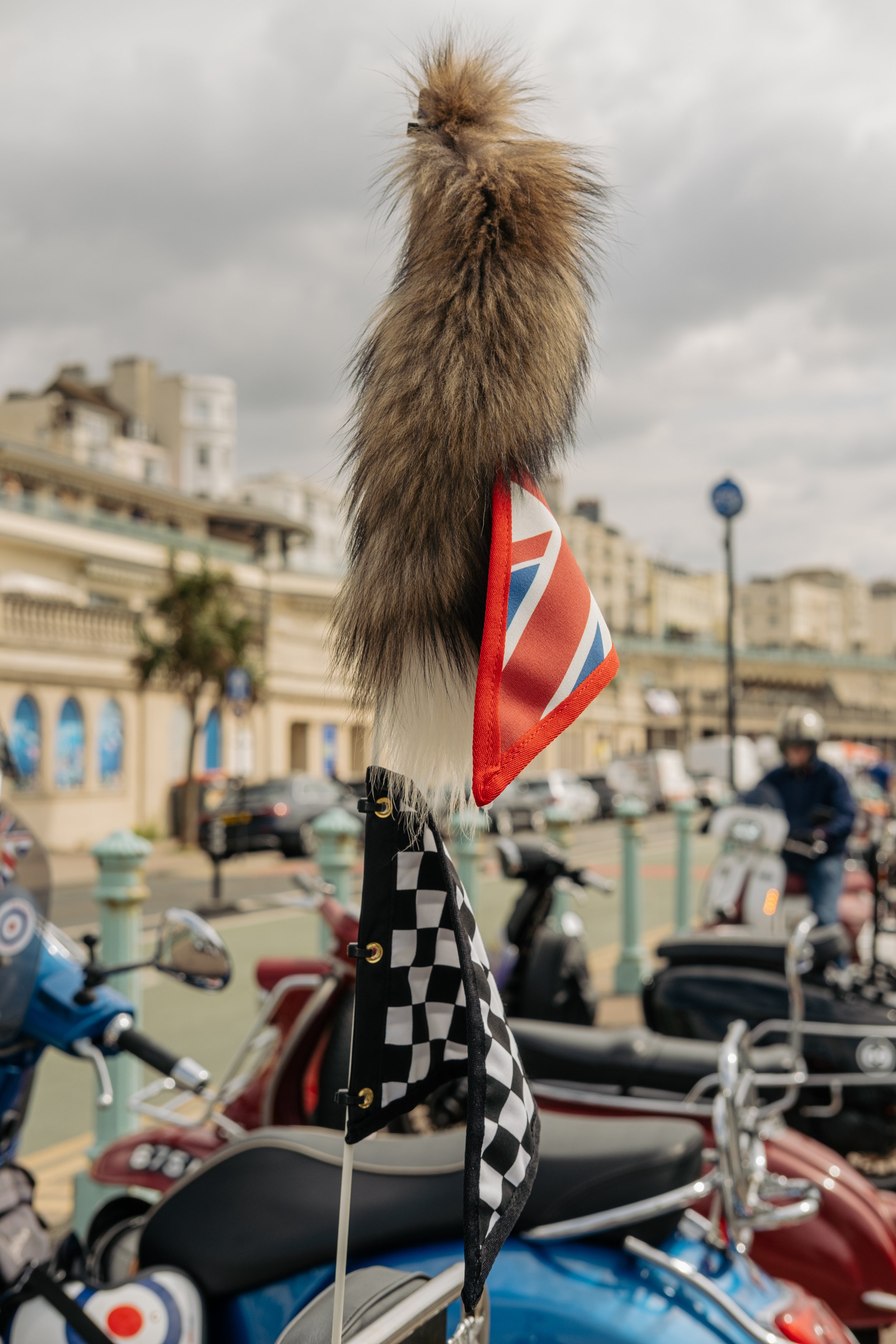 A racing flag and a fox tail accessory attached to a bike handlebar in an outdoor parking area with motorcycles and people in the background.