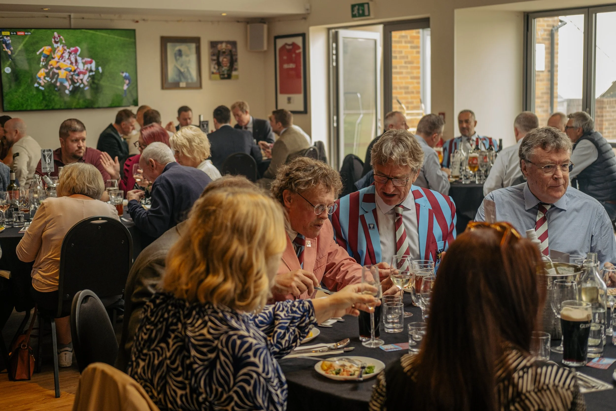 People gathered at a banquet table, some wearing striped blazers, watching a rugby game on a large screen in a banquet hall.