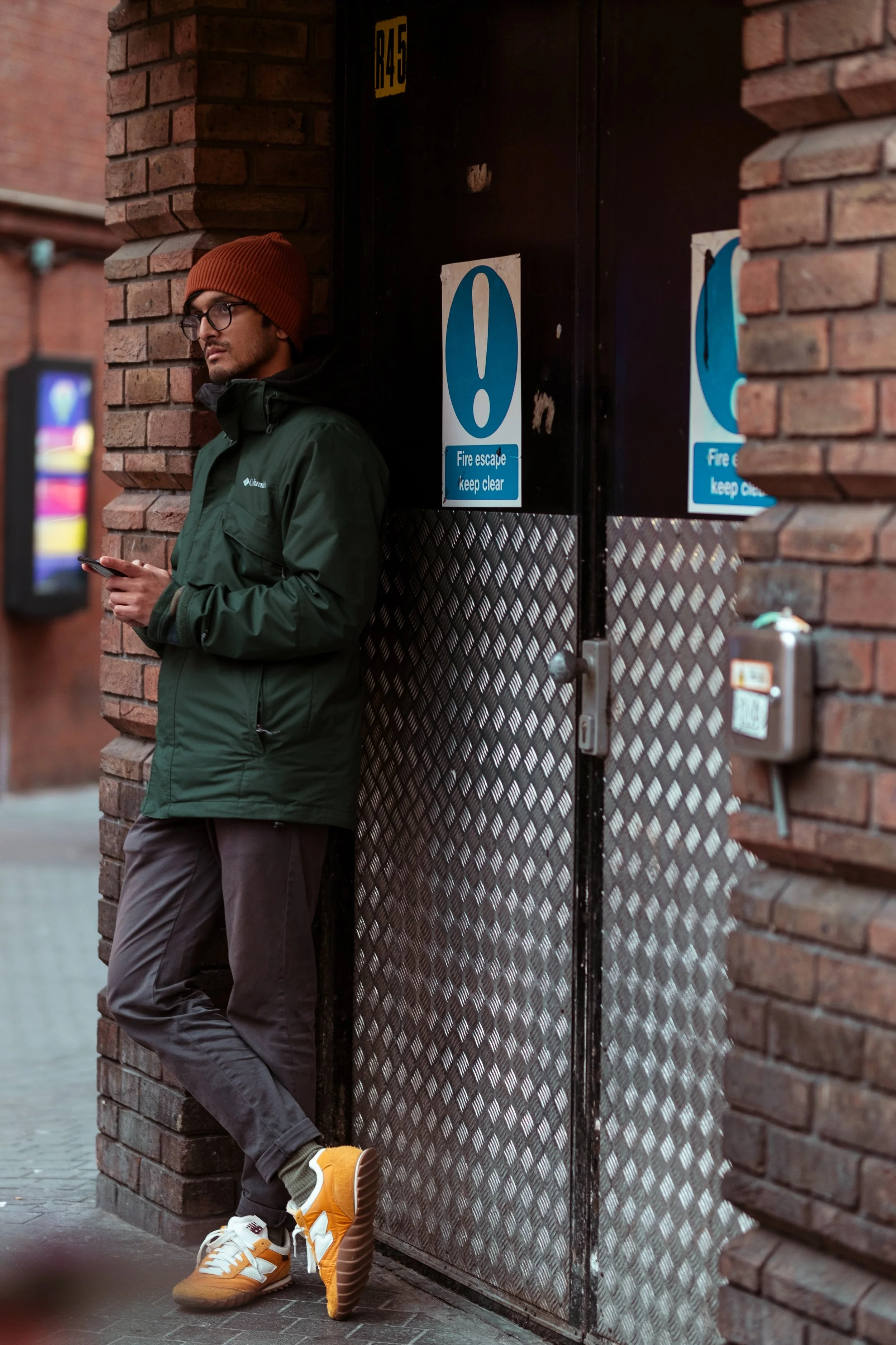 A man in a green jacket, orange beanie, and yellow sneakers leaning against a brick wall while looking at his phone.