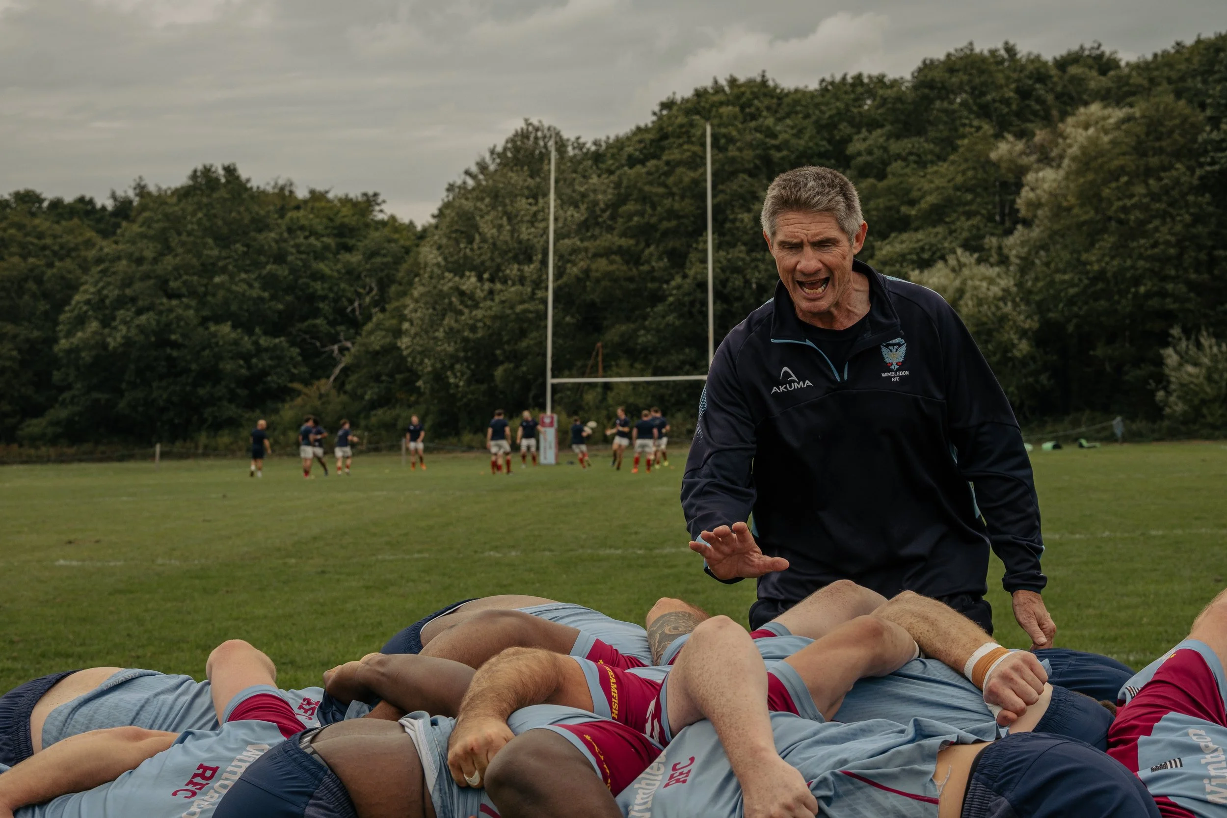 Rugby coach shouting and gesturing during a scrum on a rugby field with players and goal posts, green trees and overcast sky in the background.