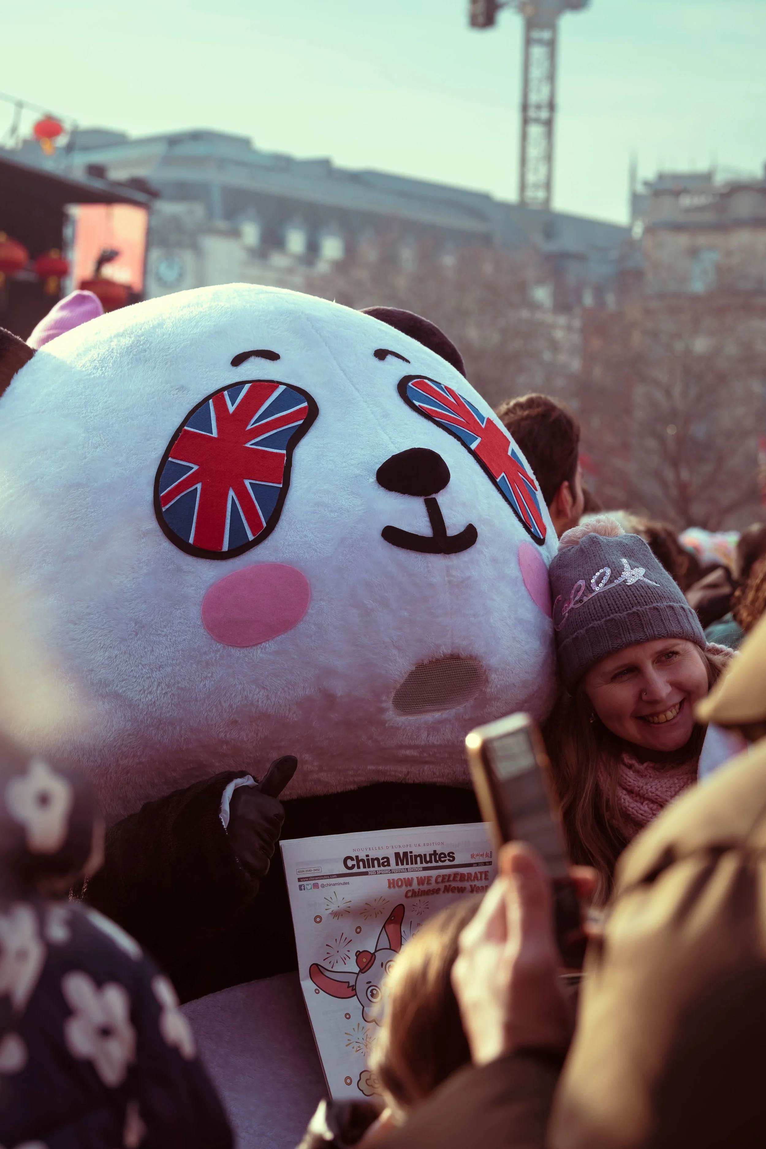 A person dressed in a large panda mascot costume with eyes featuring Union Jack flags, surrounded by a smiling crowd celebrating a Chinese New Year event.