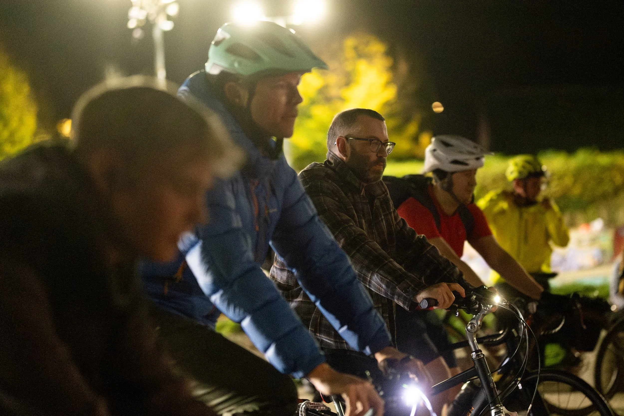 A group of people riding bicycles at night, wearing helmets and outdoor clothing, illuminated by bike lights.