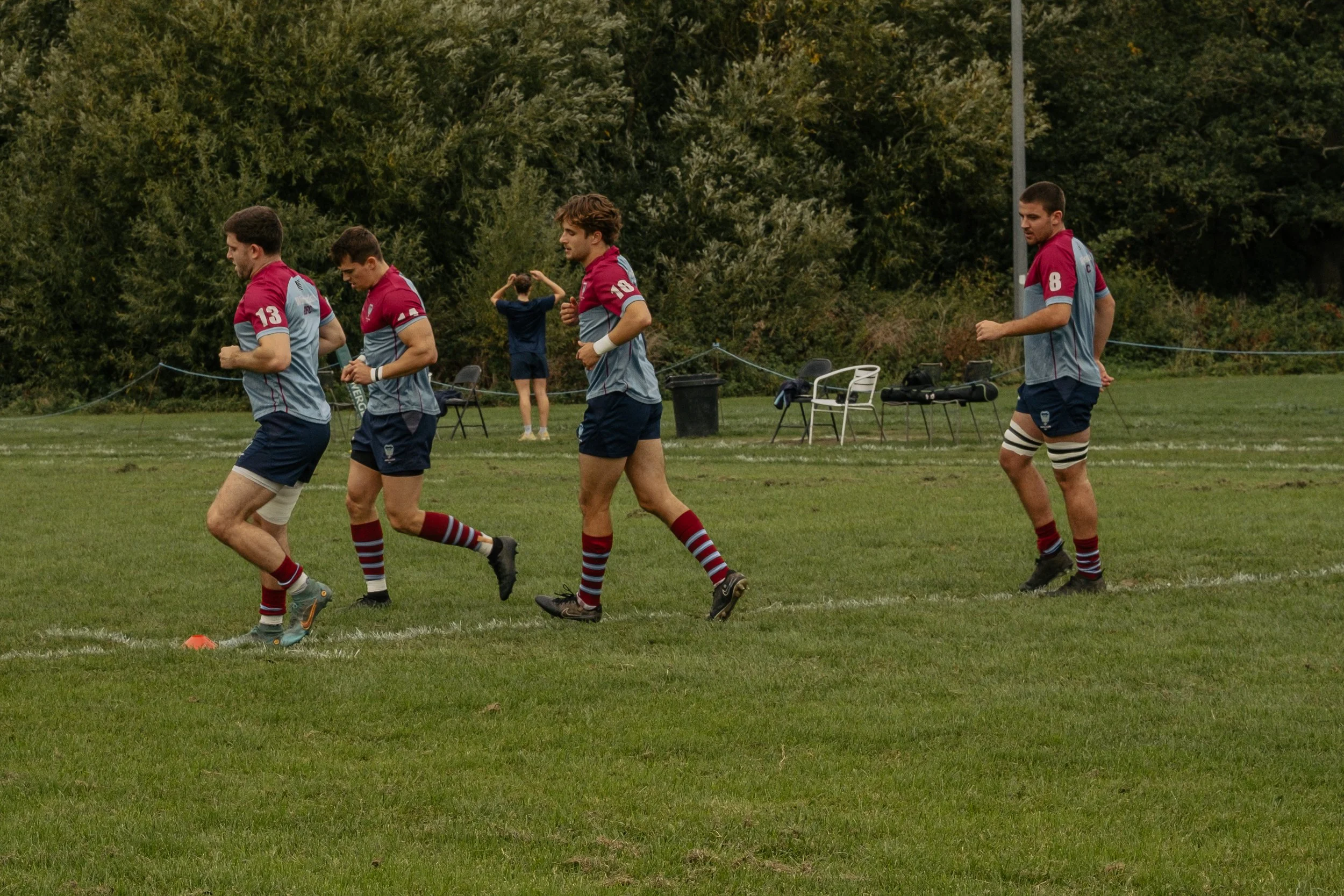 Rugby players warming up on a grassy field, wearing matching gray and maroon uniforms, with trees in the background.