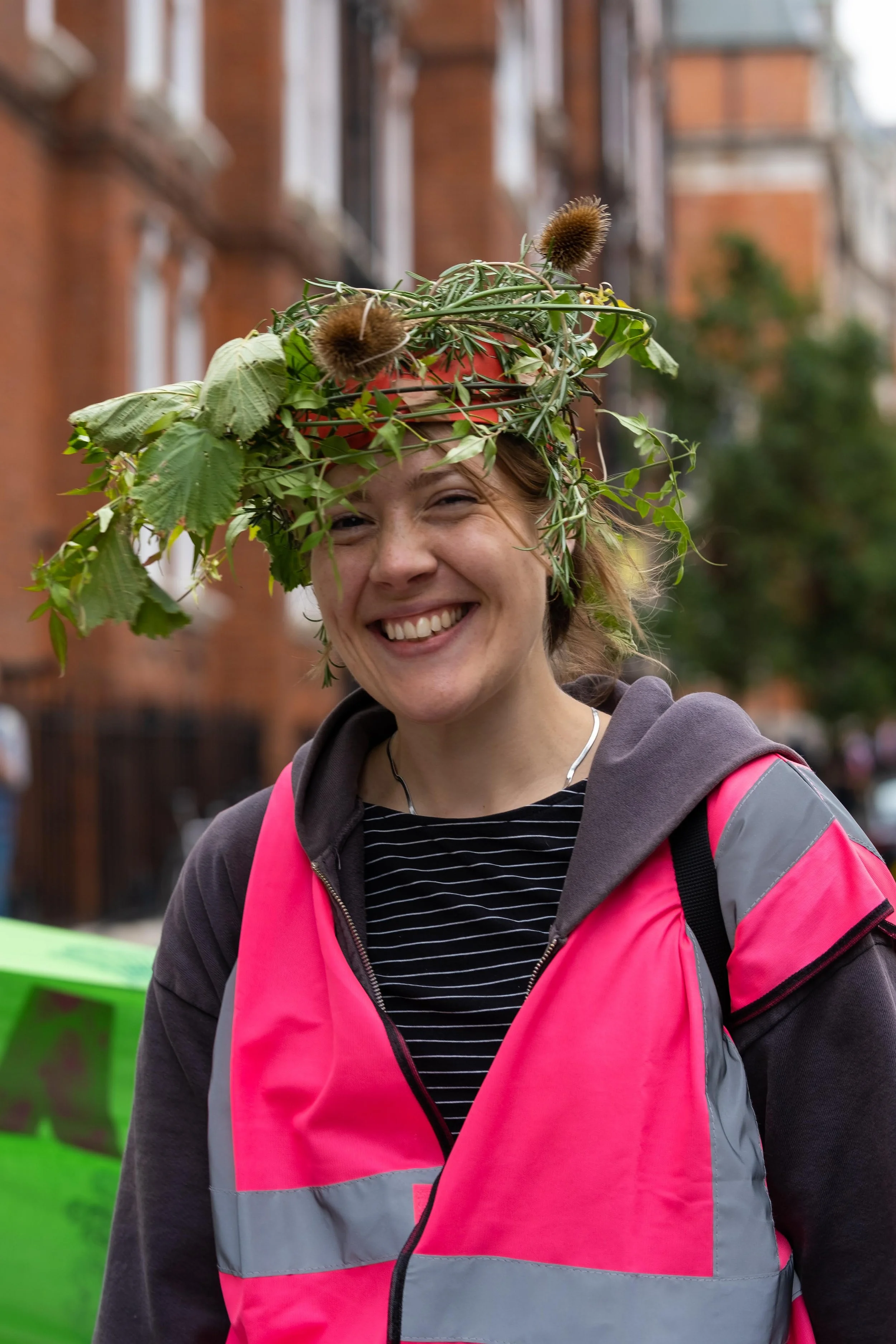 A woman smiling wearing a pink safety vest with a crown made of leaves, twigs, and burrs on her head, outdoors with brick buildings in the background.