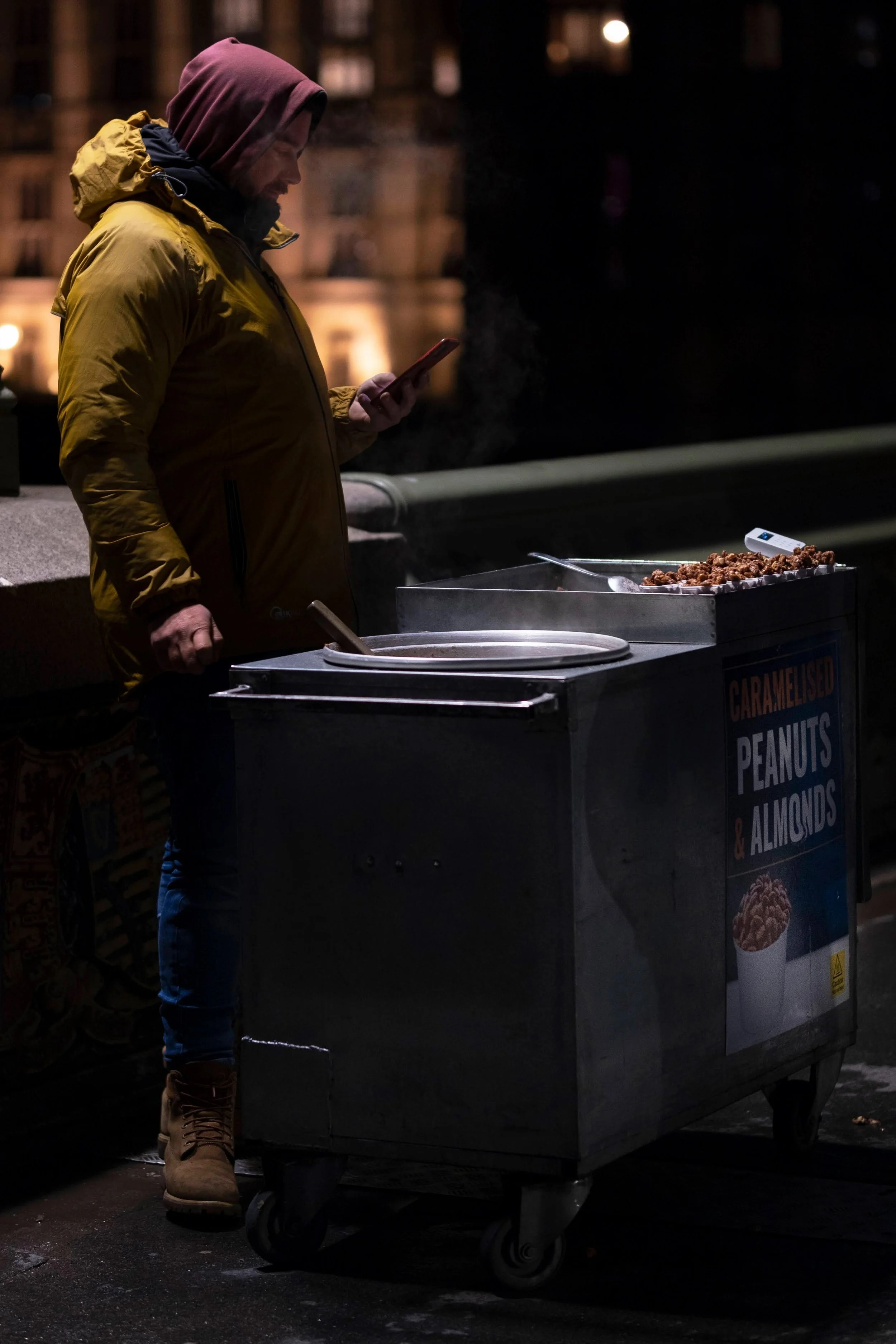 A person in a yellow jacket and pink hoodie standing at a street food cart at night, looking at their phone. The cart displays a sign for caramelized peanuts and almonds, with some food items on top.