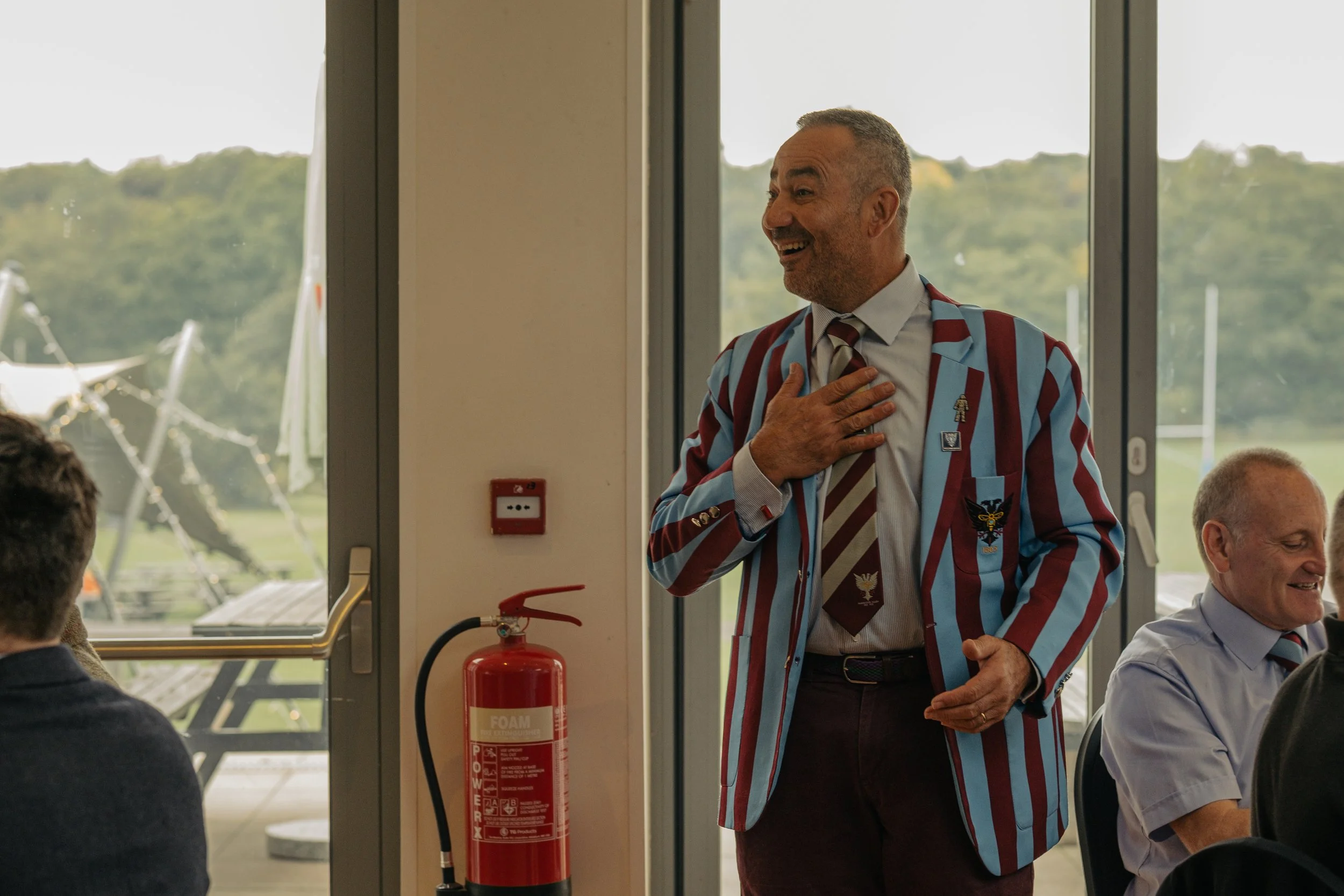 A man in a colorful striped blazer with patches, a dress shirt, and a tie with a pattern, stands indoors with his hand over his chest, smiling and looking to the right. Other people are seated nearby, and a large window behind him shows an outdoor sc