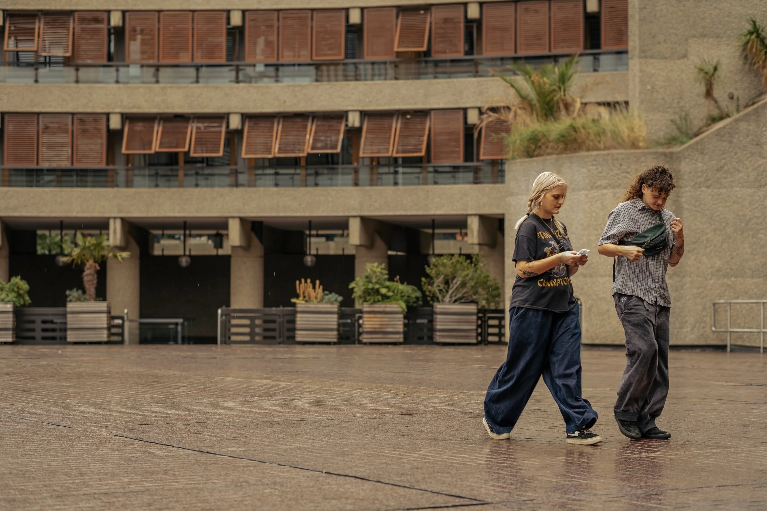 Two young women walking on wet pavement in front of a modern building with plants and open windows.