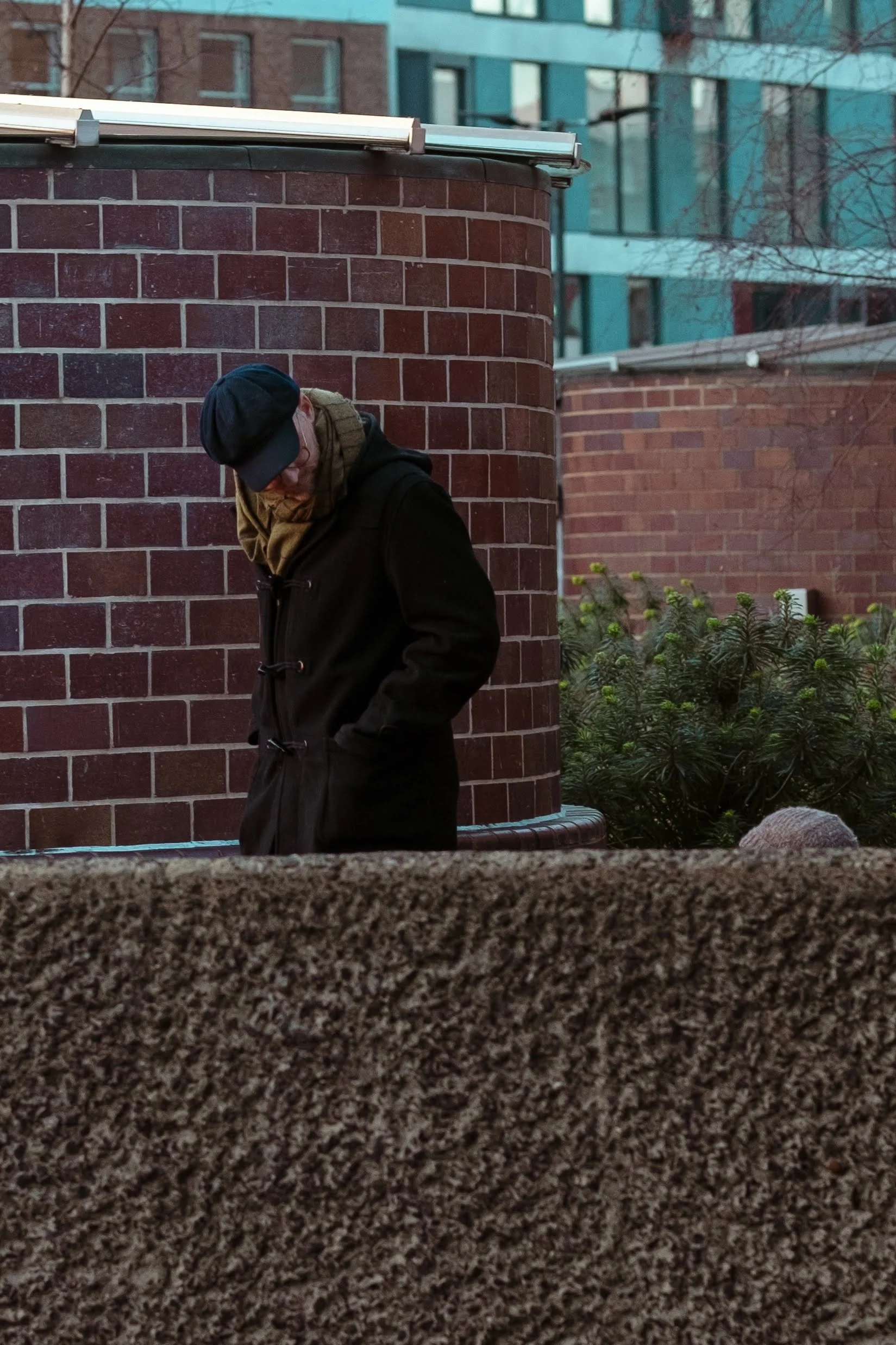 A person in a black coat, hat, and glasses standing outdoors near a red brick wall, with some greenery and modern buildings in the background.