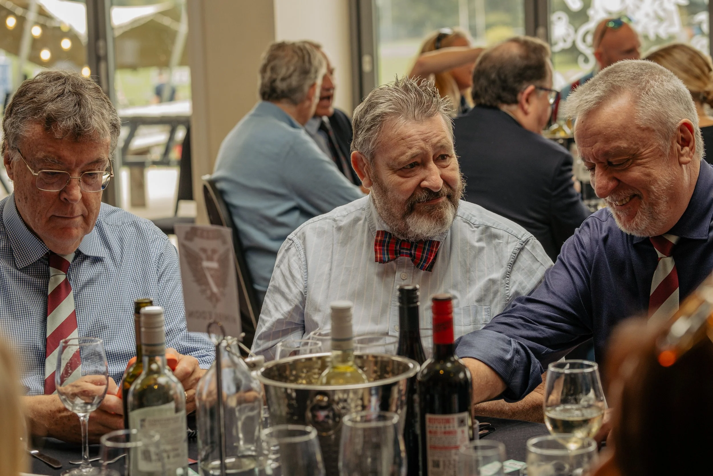 A group of older men at a table during a social event, with bottles, glasses, and utensils on the table, inside a well-lit venue with large windows.
