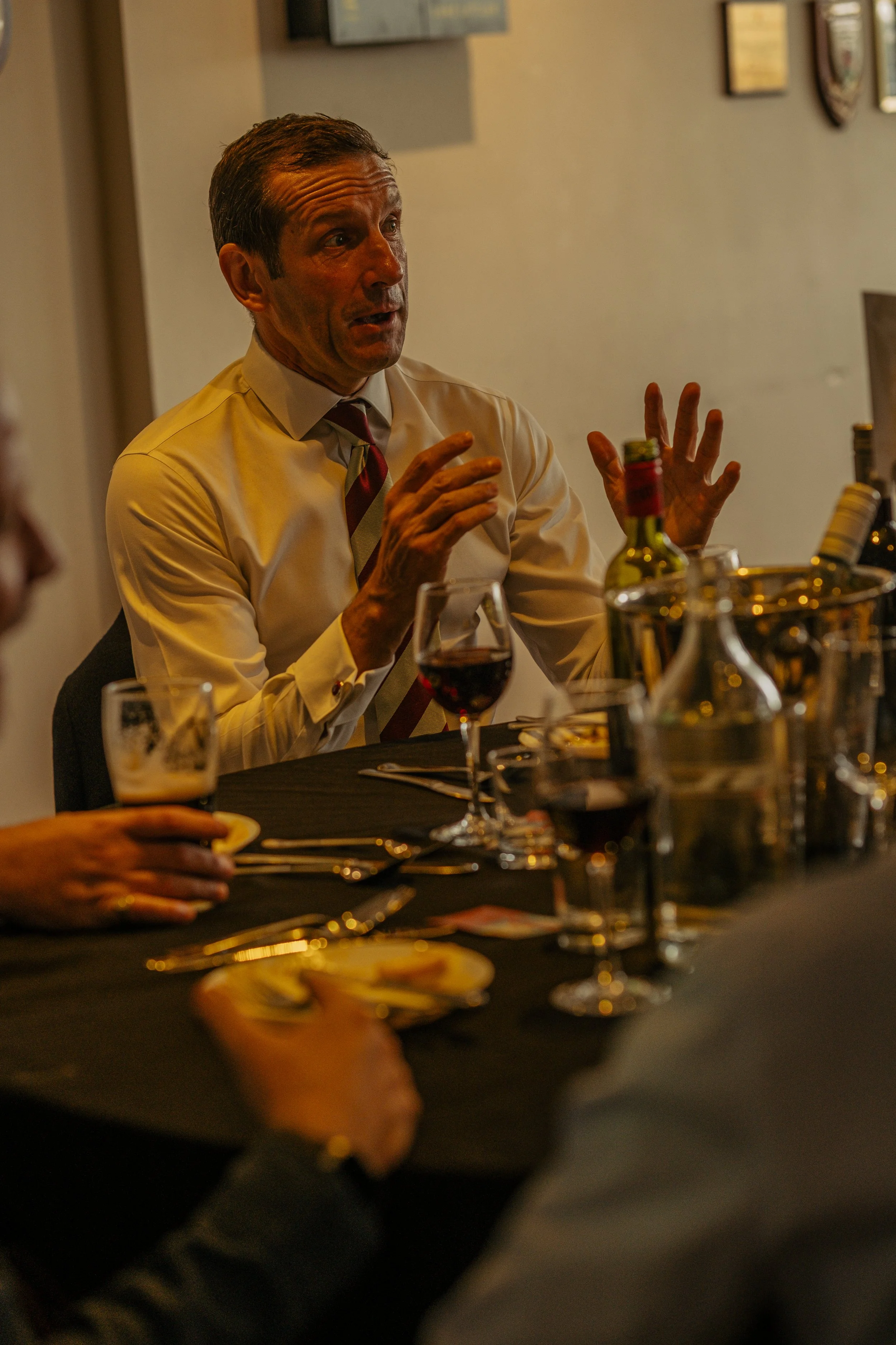 A man in a white shirt and striped tie speaking at a dinner table with wine glasses, plates, and bottles on the table, during a formal event.