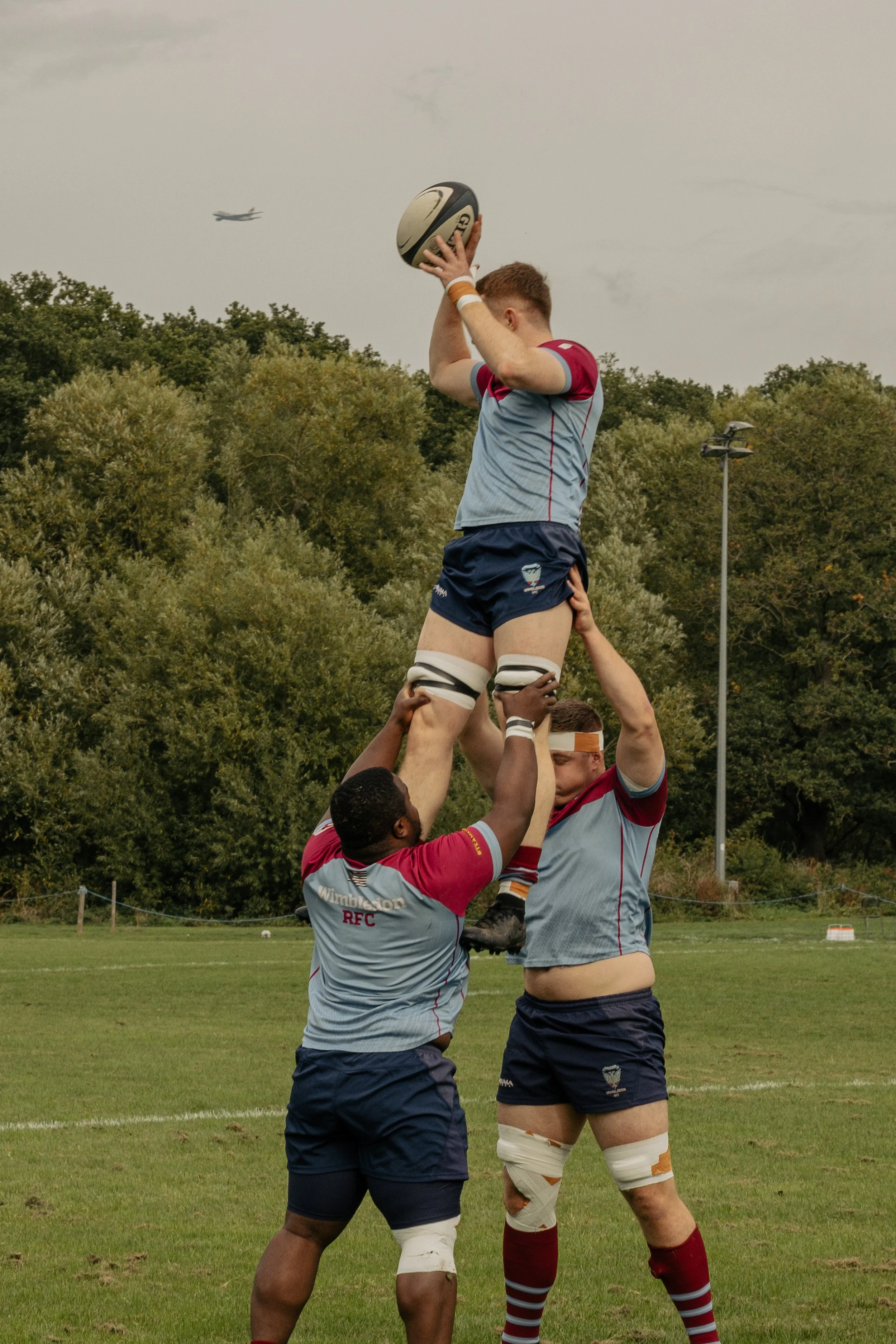 Rugby players perform a lineout, lifting a player to catch a rugby ball during a match on a grassy field with trees and an airplane in the background.