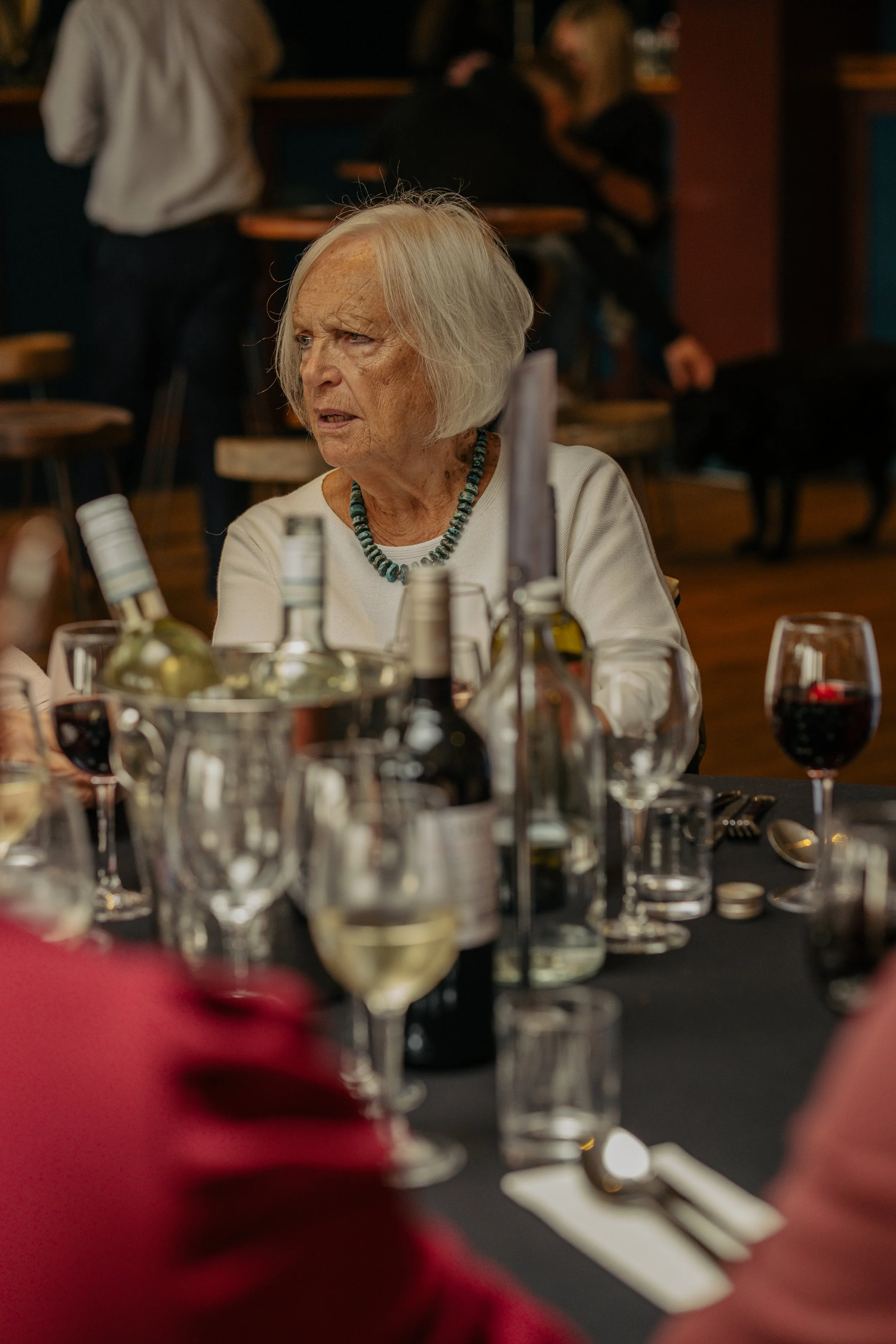 An elderly woman with gray hair, wearing a white top and a beaded necklace, sitting at a table with wine bottles and glasses, looking to the side with a surprised or concerned expression at a social gathering.