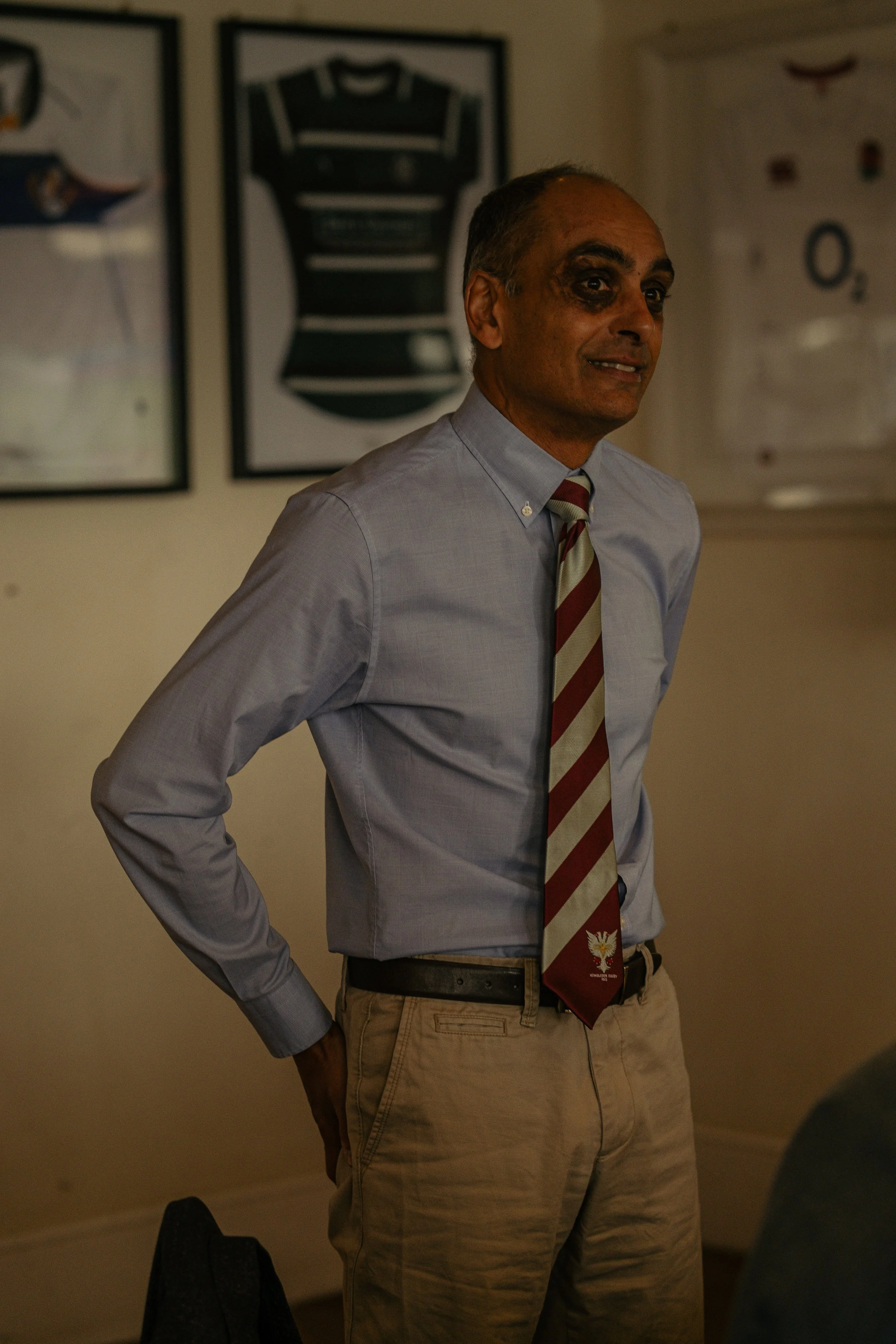 A man with dark hair and brown skin wearing a light blue dress shirt, a red and beige striped tie, and khaki pants, standing with his left hand on his hip in a room with framed sports jerseys on the wall.